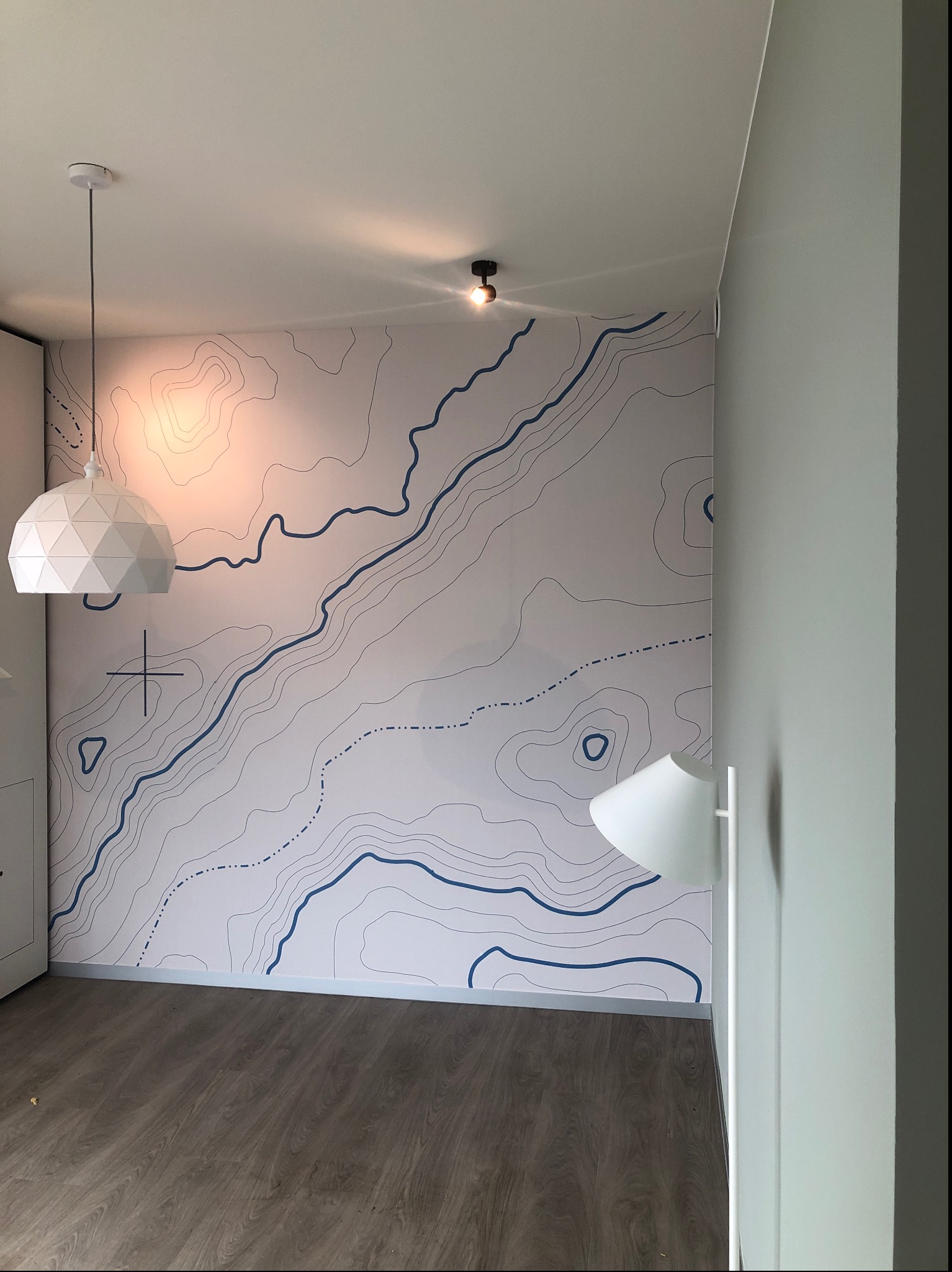 Ściana w pokoju z tapetą przedstawiającą mapę topograficzną w odcieniach bieli i niebieskiego, oświetlona przez białą lampę wiszącą o geometrycznym kształcie i lampę sufitową, z widoczną podłogą...