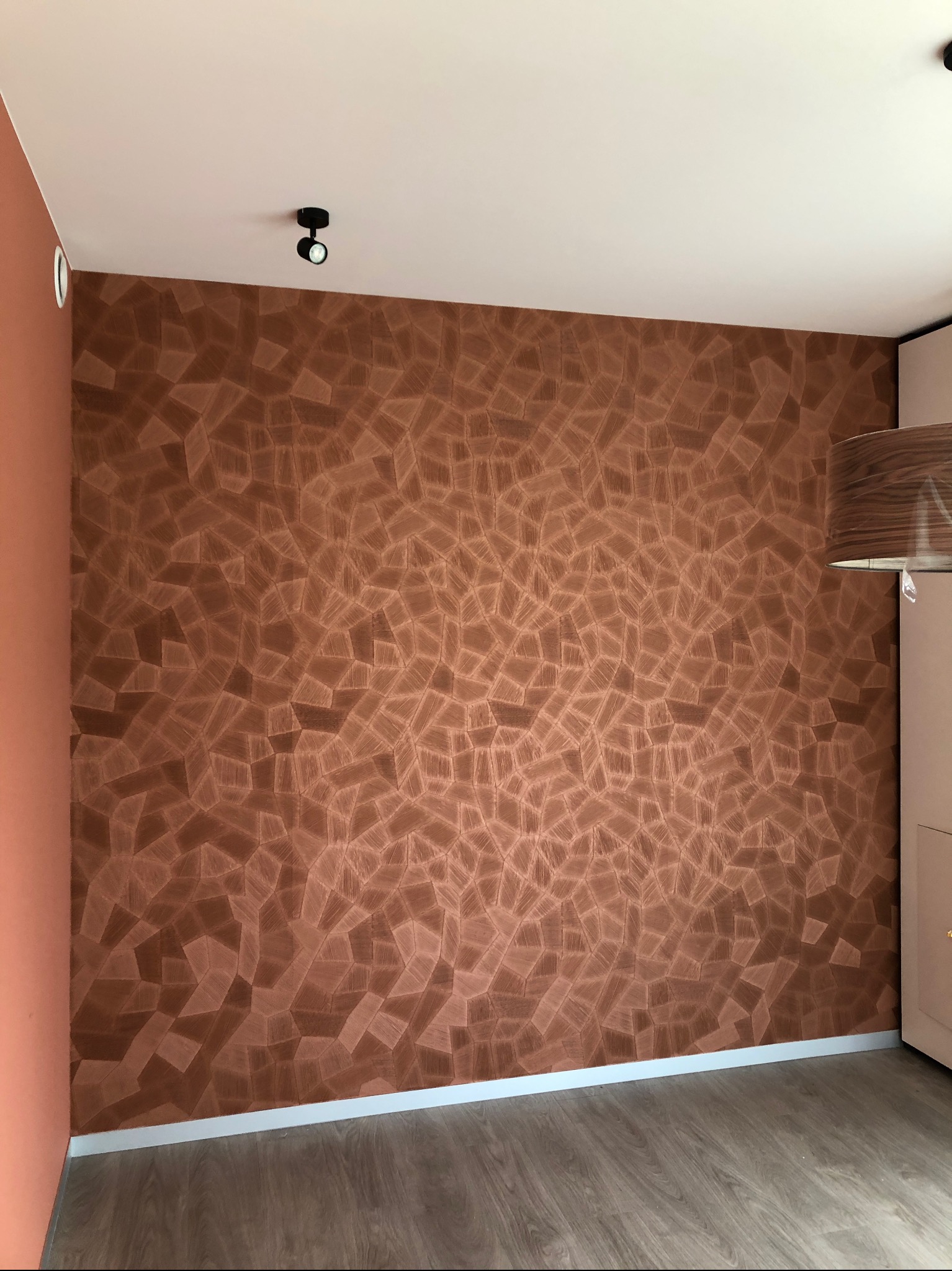 Ściana z dekoracyjną, brązową fakturą przypominającą mozaikę z drewnianych elementów, oświetlona punktowym światłem, z widocznym fragmentem szafy i podłogi.