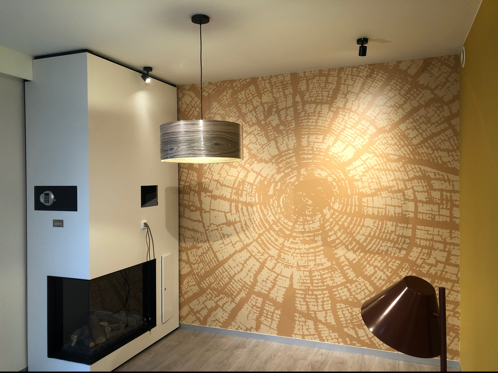 Wnętrze z dekoracyjną tapetą imitującą przekrój pnia drzewa w Pile, widoczny kominek z szybą i lampa wisząca z abażurem o fakturze drewna.