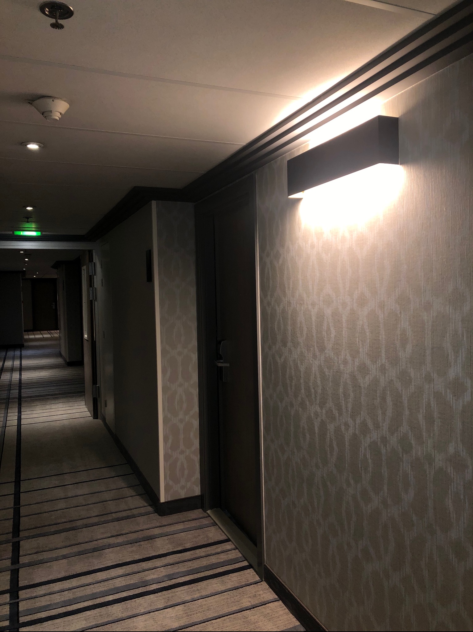 Korytarz hotelowy z tapetą w geometryczny wzór i nowoczesnym oświetleniem ściennym, widoczny fragment wykładziny dywanowej w pasy.