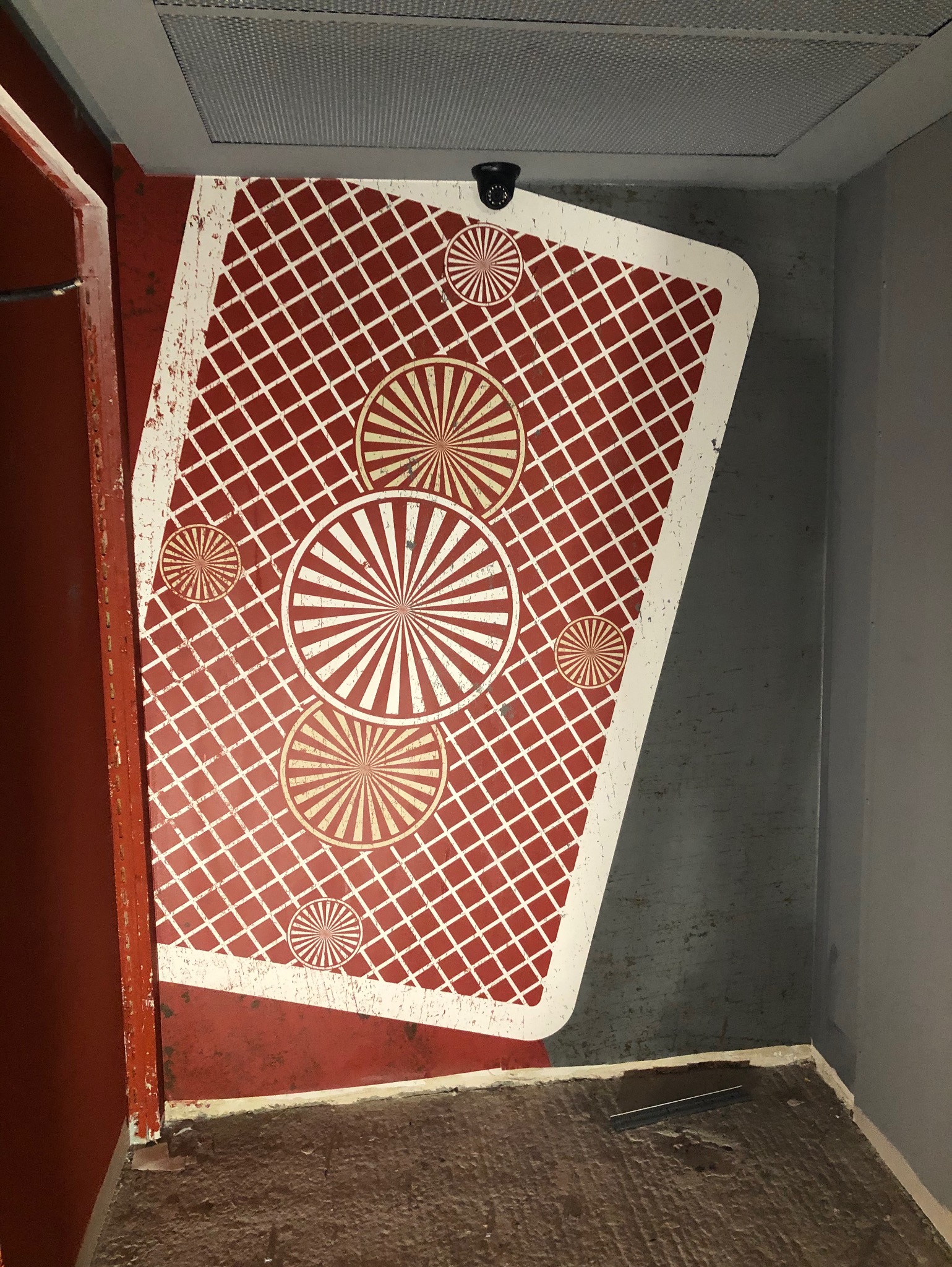 Ściana z dekoracyjnym wzorem w kratę i okręgi, przypominającym kartę do gry, w pomieszczeniu z widoczną instalacją sufitową i kamerą przemysłową.