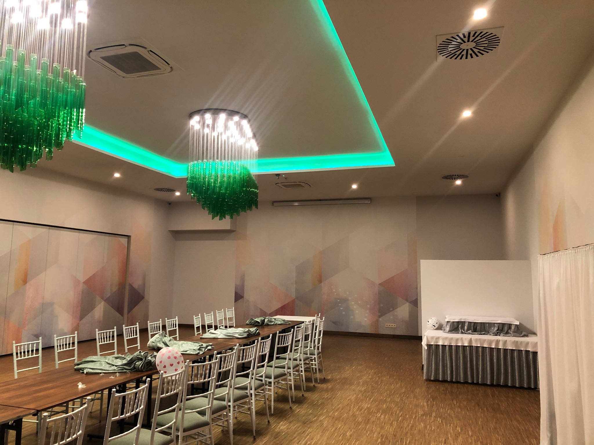 Sala konferencyjna z długim stołem przygotowanym do dekoracji, białe krzesła, zielone akcenty oświetleniowe i geometryczny wzór na ścianie
