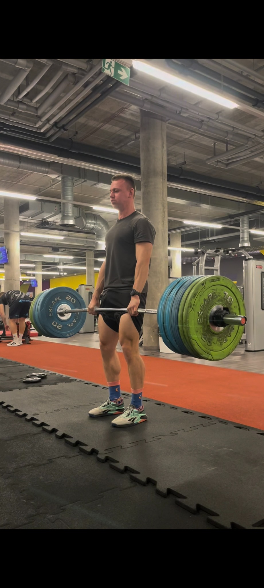 Mężczyzna w szarym t-shircie i czarnych spodenkach wykonuje martwy ciąg z dużym obciążeniem w siłowni, widoczne kolorowe talerze obciążeniowe i charakterystyczne skarpetki.