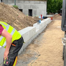 USŁUGI DROGOWE I BRUKARSKIE RADOSŁAW WOJTOWICZ - Montaż prefabrykowanych betonowych elementów ogrodzenia na terenie budowy, widoczny pracownik w odblaskowej kamizelce i spodniach roboczych, w tle wykopana ziemia i betonowy budynek.