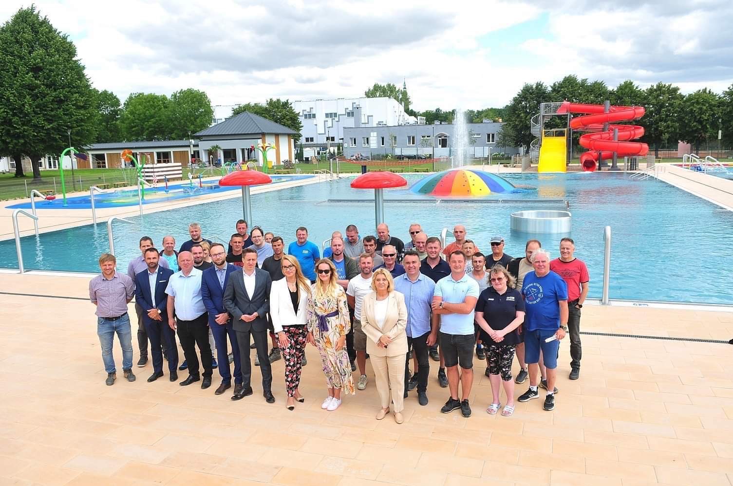 Grupa ludzi pozuje na tle nowego basenu w Świdnicy, z atrakcjami wodnymi, zjeżdżalnią i fontanną w tle.