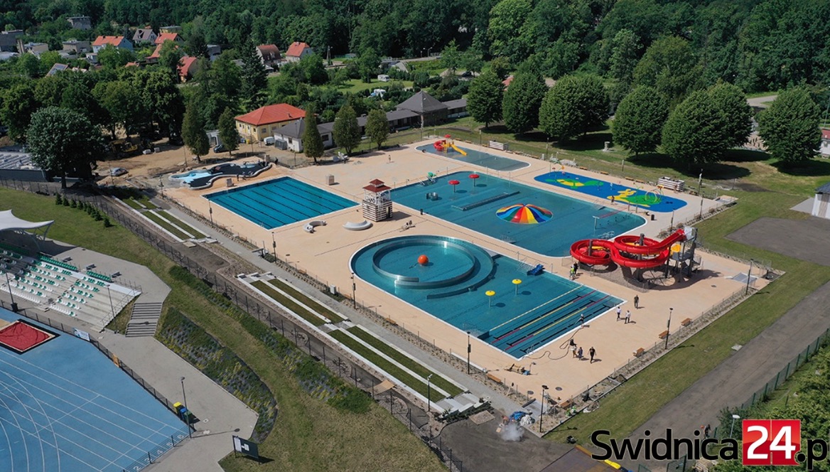 Kompleks basenów zewnętrznych w Świdnicy, widok z lotu ptaka, obejmujący basen sportowy, rekreacyjny z atrakcjami wodnymi, brodzik dla dzieci oraz czerwoną zjeżdżalnię, otoczony zielenią i budynkami.