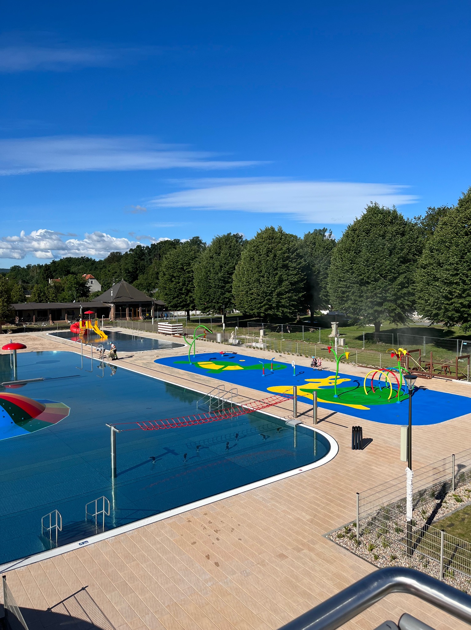 Kompleks basenów zewnętrznych z atrakcjami wodnymi dla dzieci, otoczony drzewami i budynkami rekreacyjnymi, widok z góry w słoneczny dzień.