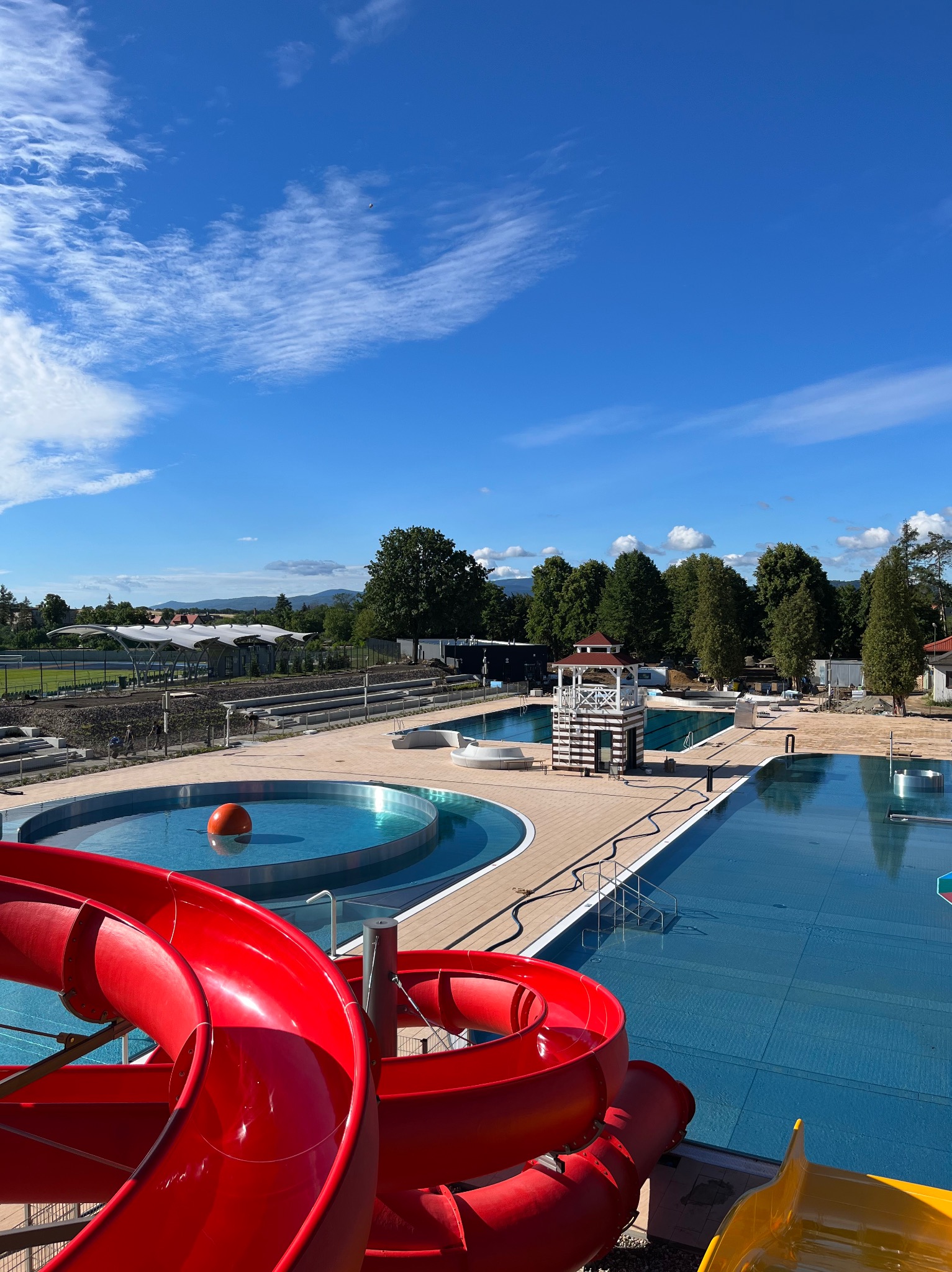 Widok z góry na kompleks basenów zewnętrznych z czerwonymi i żółtymi zjeżdżalniami, drewnianym pomostem, wieżą ratowniczą i błękitnym niebem z chmurami w tle.
