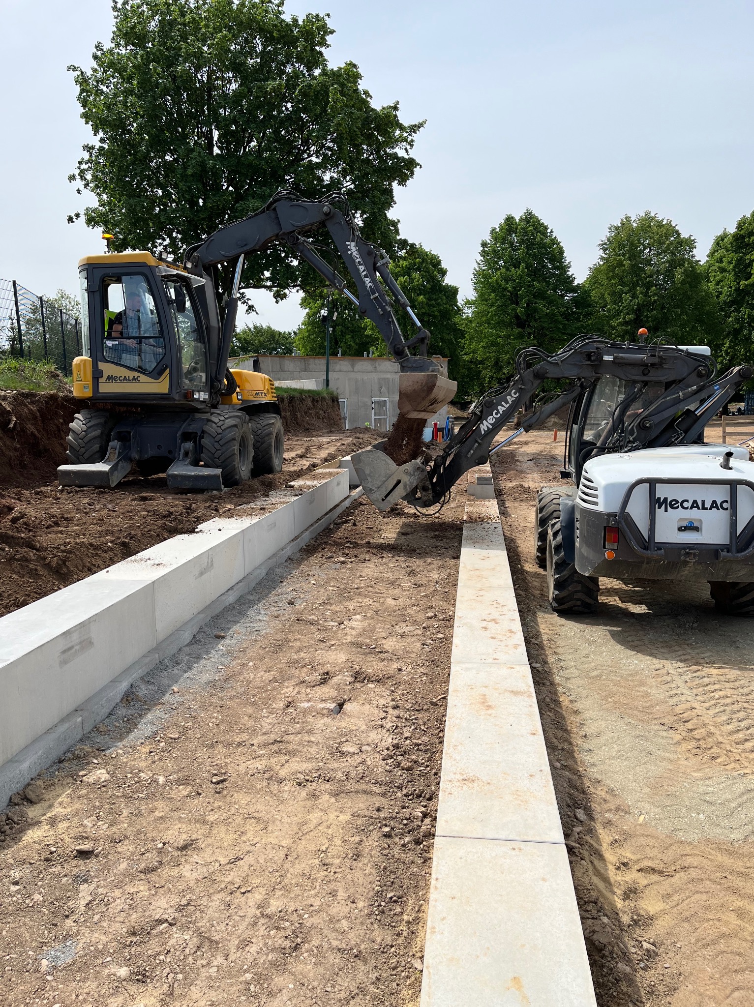 Dwa żółto-czarne koparki Mecalac pracują przy wykopie obok ułożonych betonowych krawężników na placu budowy z zielonymi drzewami w tle.