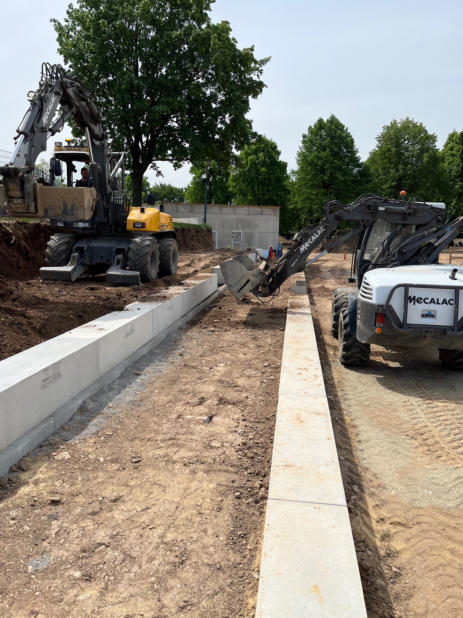 Dwa biało-szare koparki Mecalac pracują na placu budowy, układając betonowe obrzeża wzdłuż wykopu; w tle widoczne drzewa i częściowo wykopana ziemia.