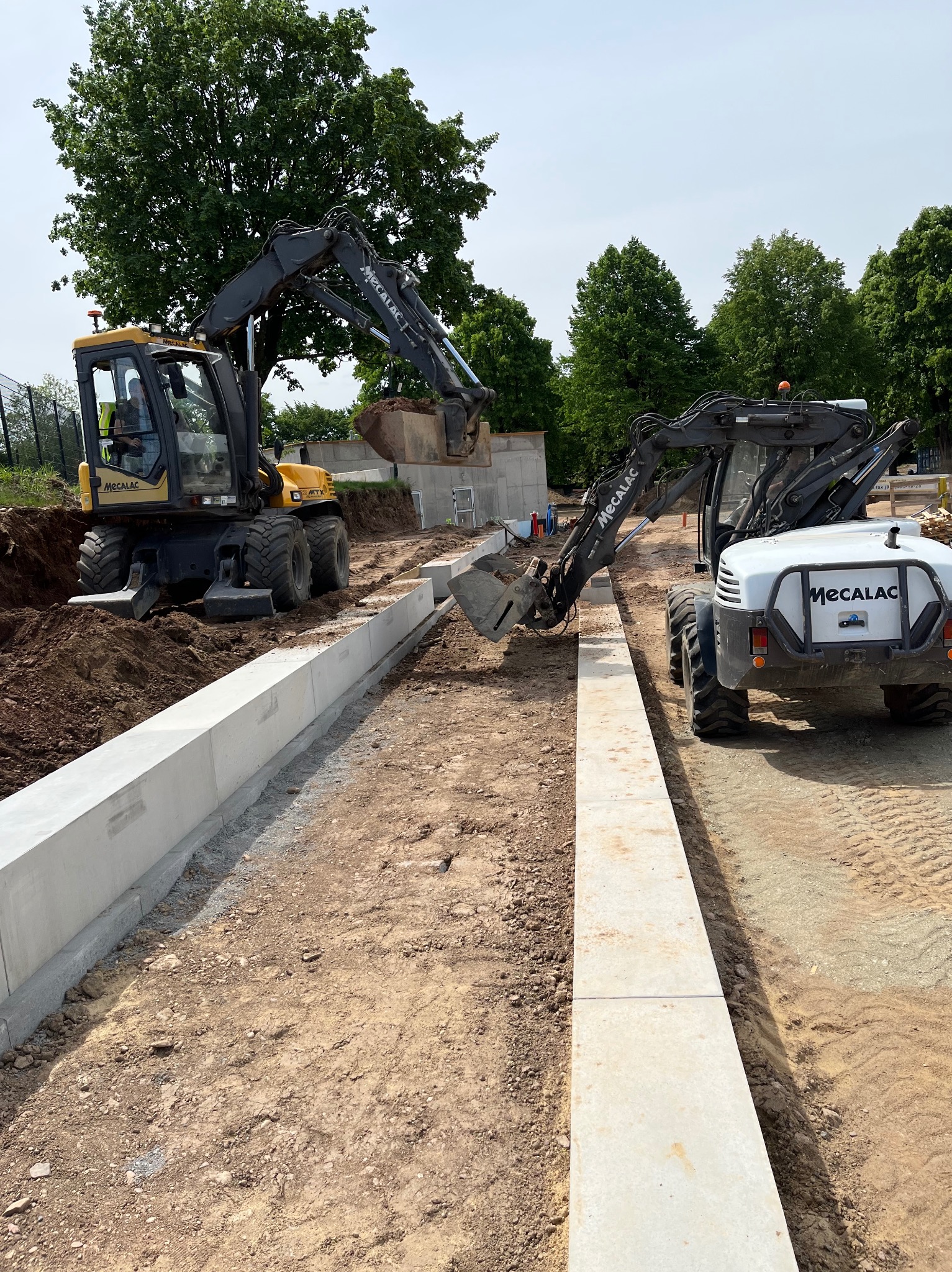 Dwa żółto-szare koparko-ładowarki Mecalac pracują przy montażu betonowych krawężników na placu budowy, z widocznym wykopem ziemnym i zielonymi drzewami w tle.