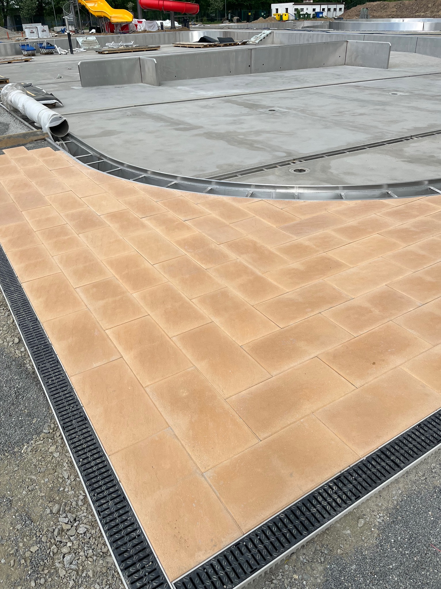 Fragment budowy basenu z ułożoną jasnobrązową kostką brukową wokół krawędzi, czarny system odprowadzania wody i widoczne elementy konstrukcyjne basenu w tle.
