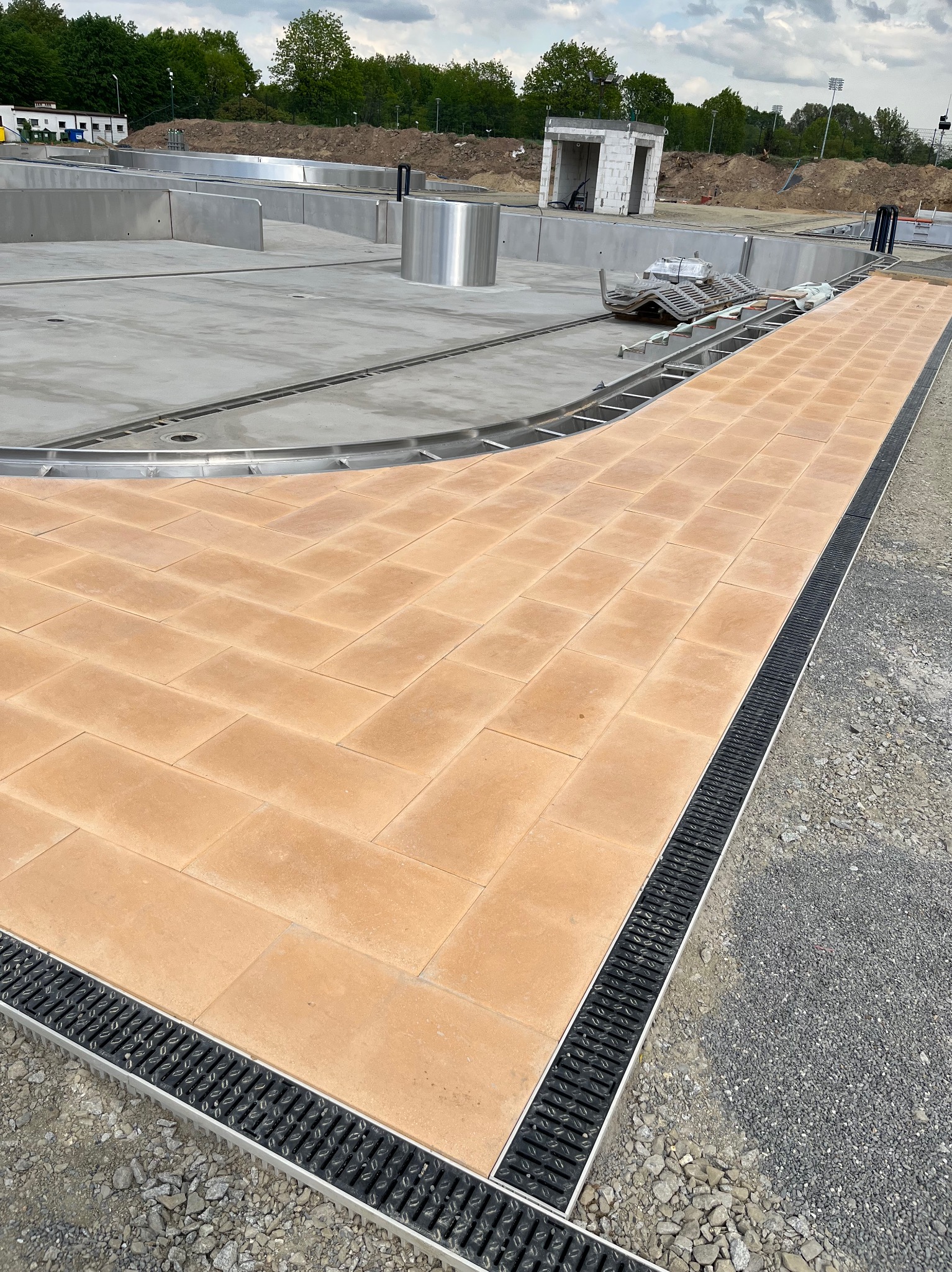 Ułożona ścieżka z kostki brukowej w jodełkę, obok wykopu pod prawdopodobny basen, z widocznym systemem odprowadzania wody w postaci czarnych kratek ściekowych.