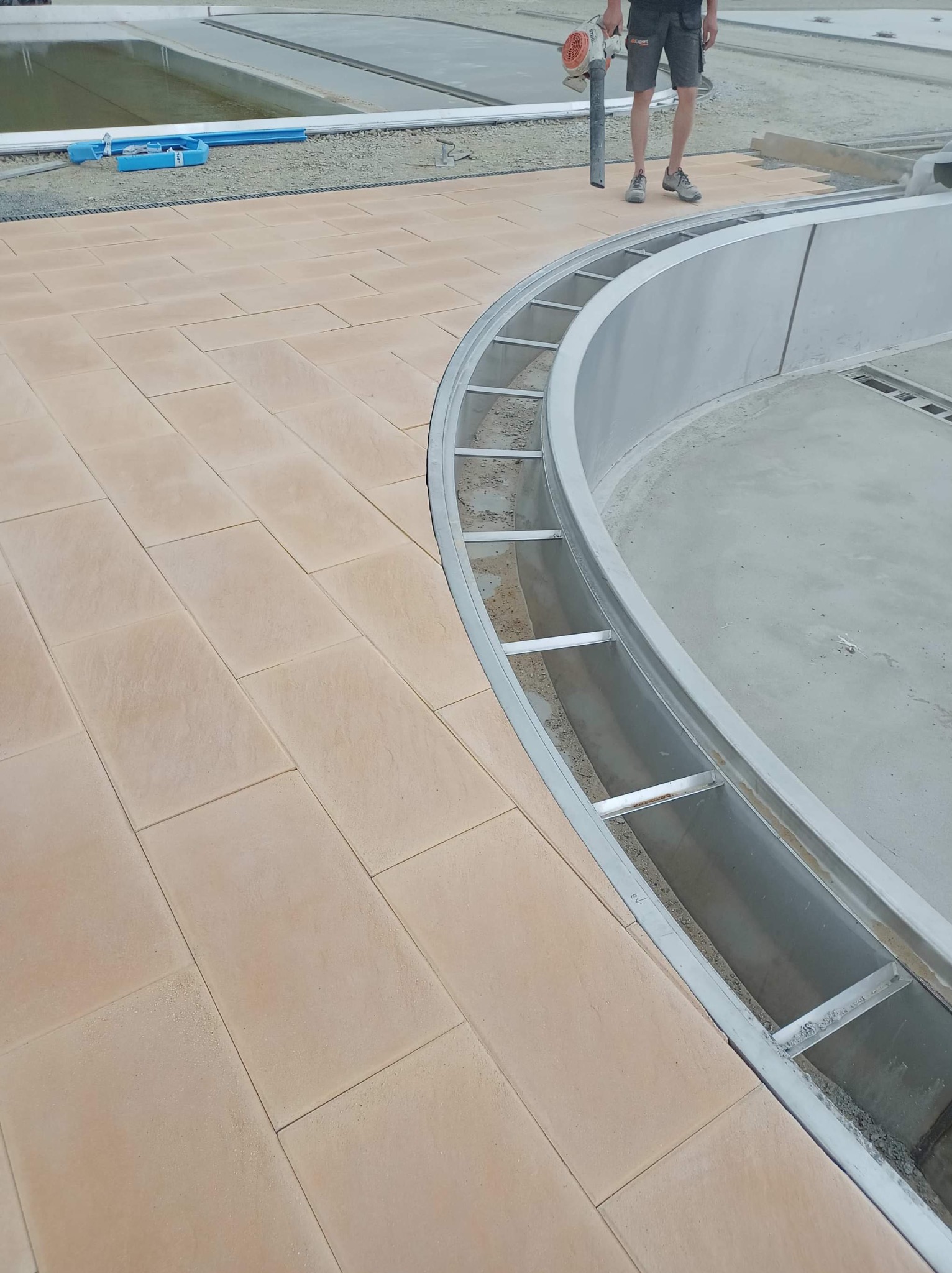 Układanie jasnej kostki brukowej wokół basenu w trakcie budowy, widoczne metalowe elementy konstrukcyjne i pracownik z dmuchawą do liści.