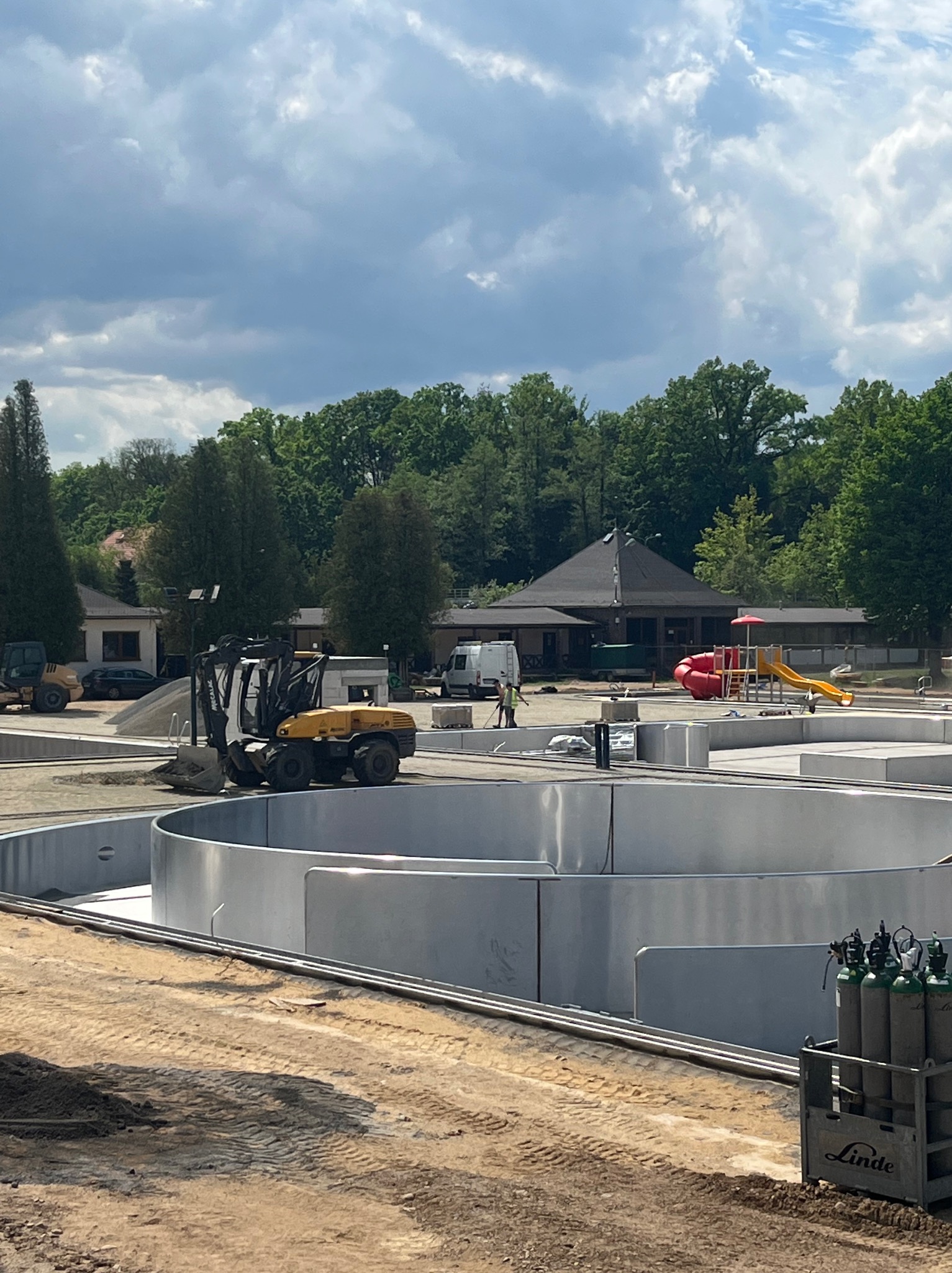 Budowa okrągłego basenu z użyciem minikoparki Mecalac i butli z gazem Linde, widok na plac budowy, budynki i plac zabaw w tle, pochmurne niebo.