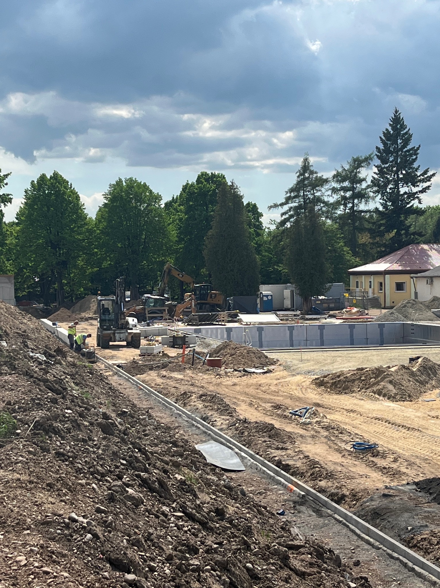 Budowa basenu w ogrodzie: robotnicy przy pracy, widoczne maszyny budowlane i wykopane fundamenty pod basen na tle zieleni i pochmurnego nieba.