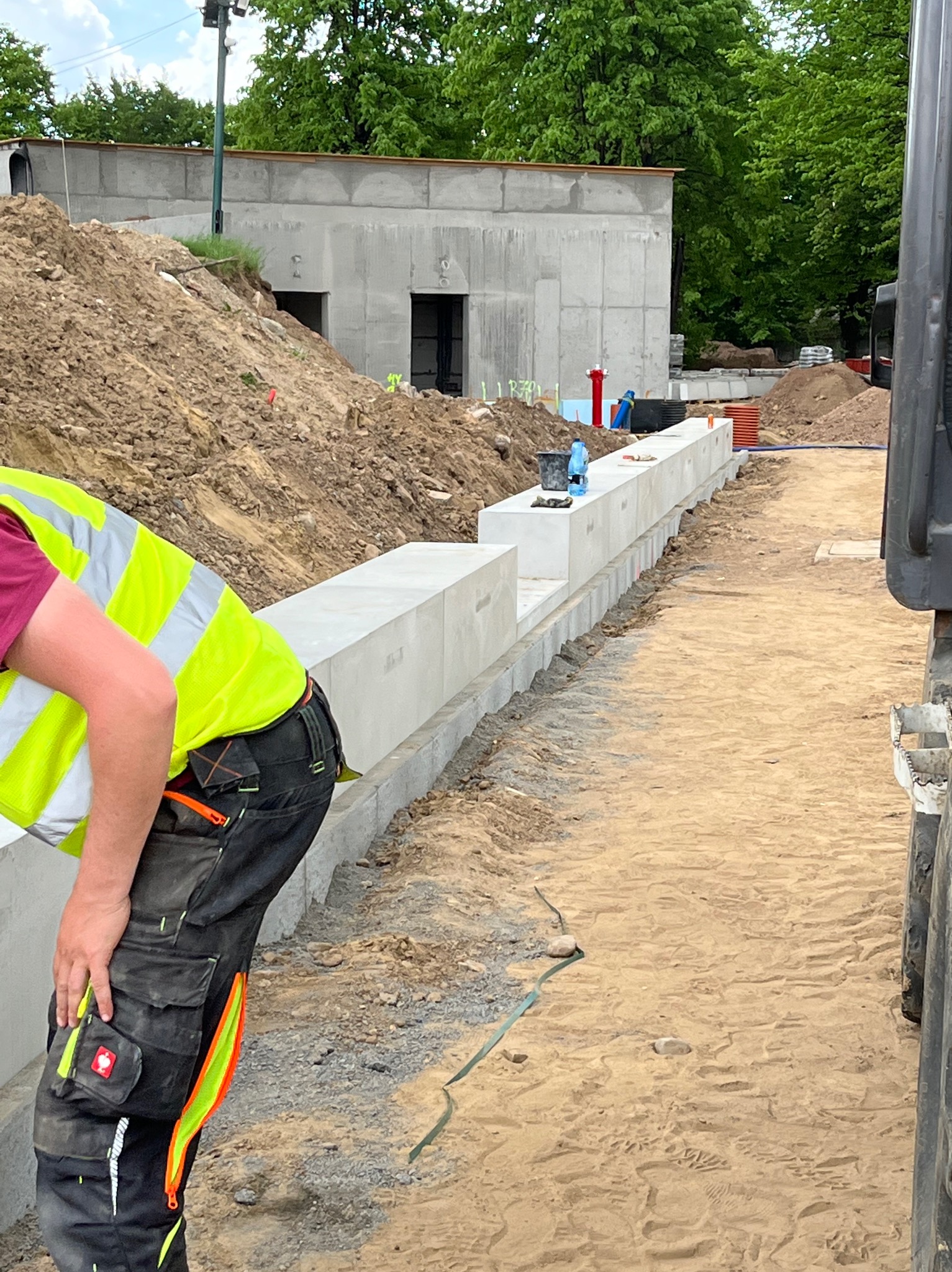 Montaż prefabrykowanych betonowych elementów ogrodzenia na terenie budowy, widoczny pracownik w odblaskowej kamizelce i spodniach roboczych, w tle wykopana ziemia i betonowy budynek.