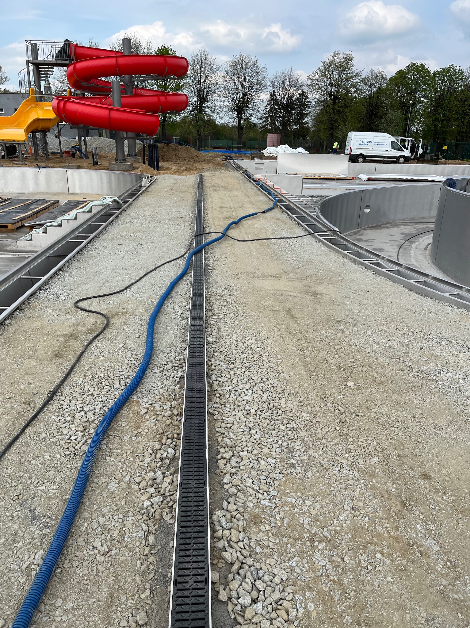 Budowa basenu w Świdnicy: widok na plac budowy z zamontowanym odpływem liniowym, w tle zjeżdżalnie i samochód dostawczy firmy Berndorf Bäderbau.