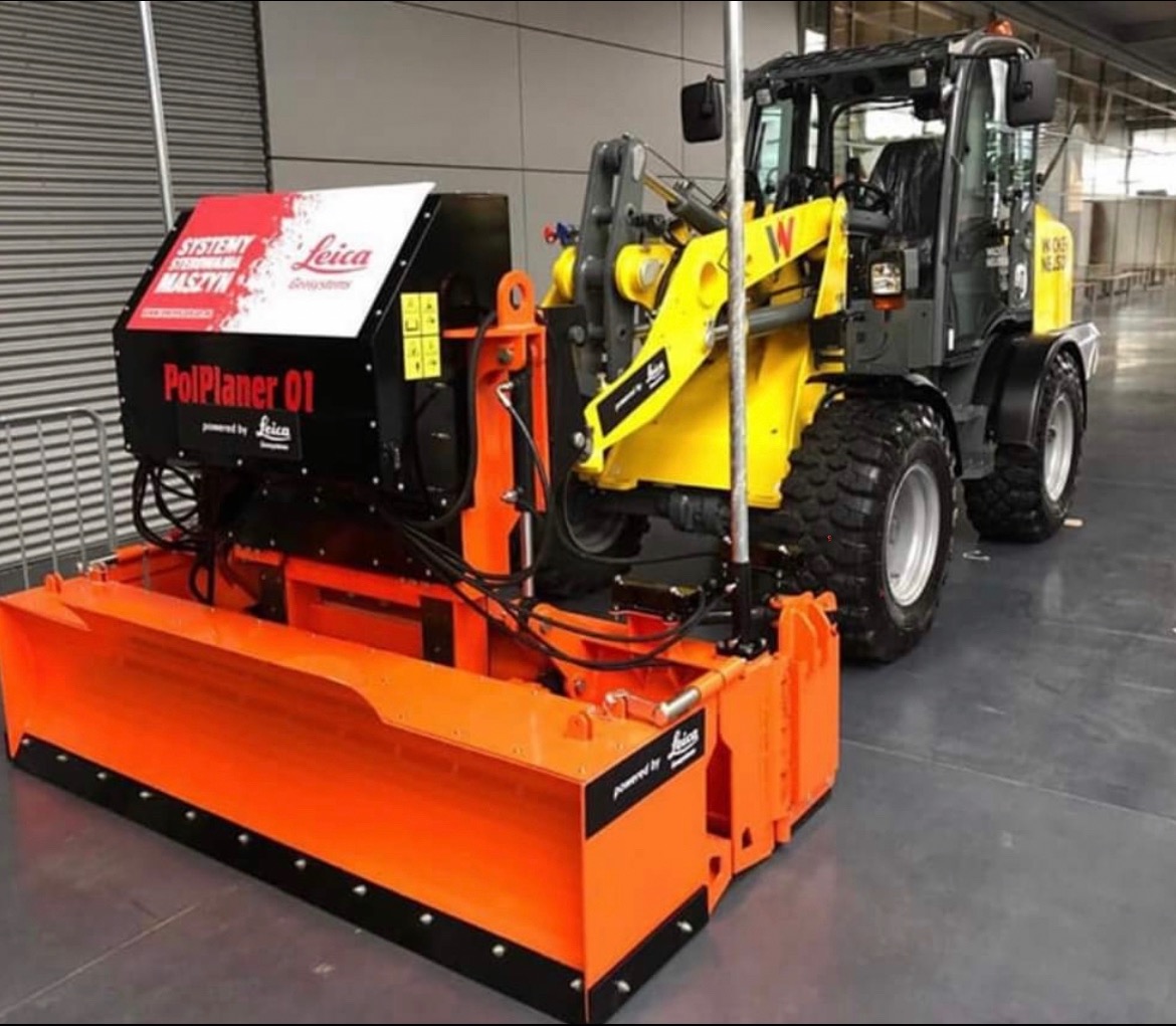 Żółto-szara ładowarka Wacker Neuson z pomarańczowym osprzętem PolPlaner 01 firmy Leica, prezentowana w hali.