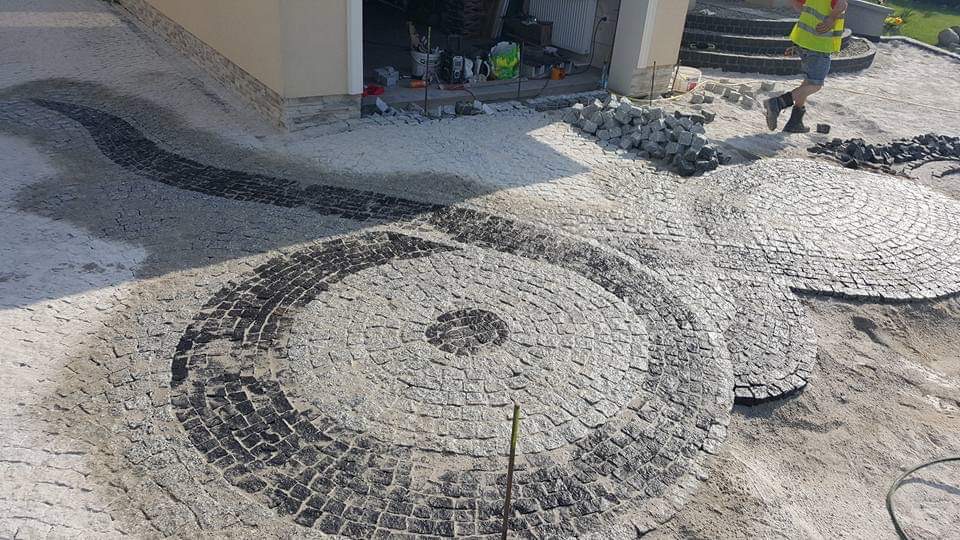 Niedokończona nawierzchnia z kostki brukowej w kształcie okręgów i wijącej się ścieżki, widoczne narzędzia i zapasy kostki, pracownik w odblaskowej kamizelce w tle.