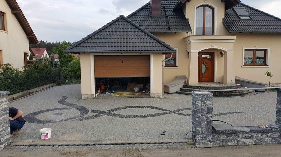 Nowoczesny podjazd z kostki brukowej z dekoracyjnym wzorem wijącej się wstęgi, częściowo ukończony, z widocznym fragmentem ogrodzenia i pracownikiem w trakcie realizacji, dom w tle.