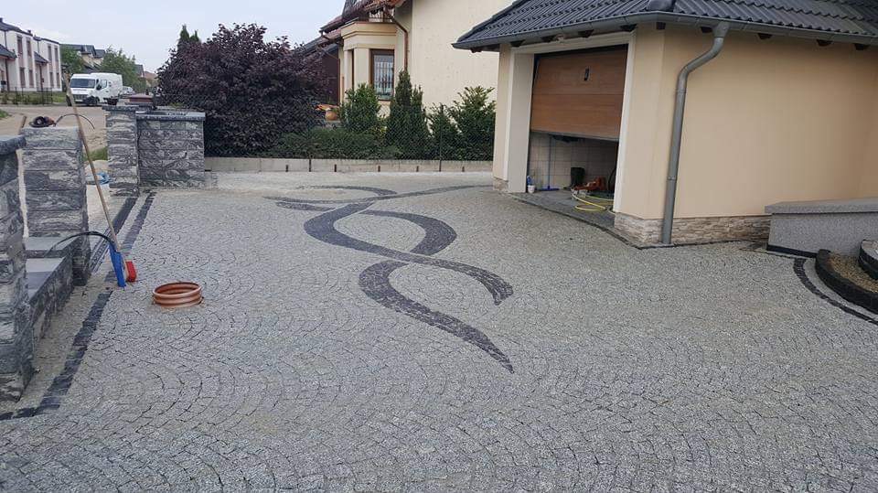 Podjazd z jasnoszarej kostki granitowej z dekoracyjnym, ciemnoszarym wzorem w kształcie podwójnej spirali, widok na garaż i fragment posesji.