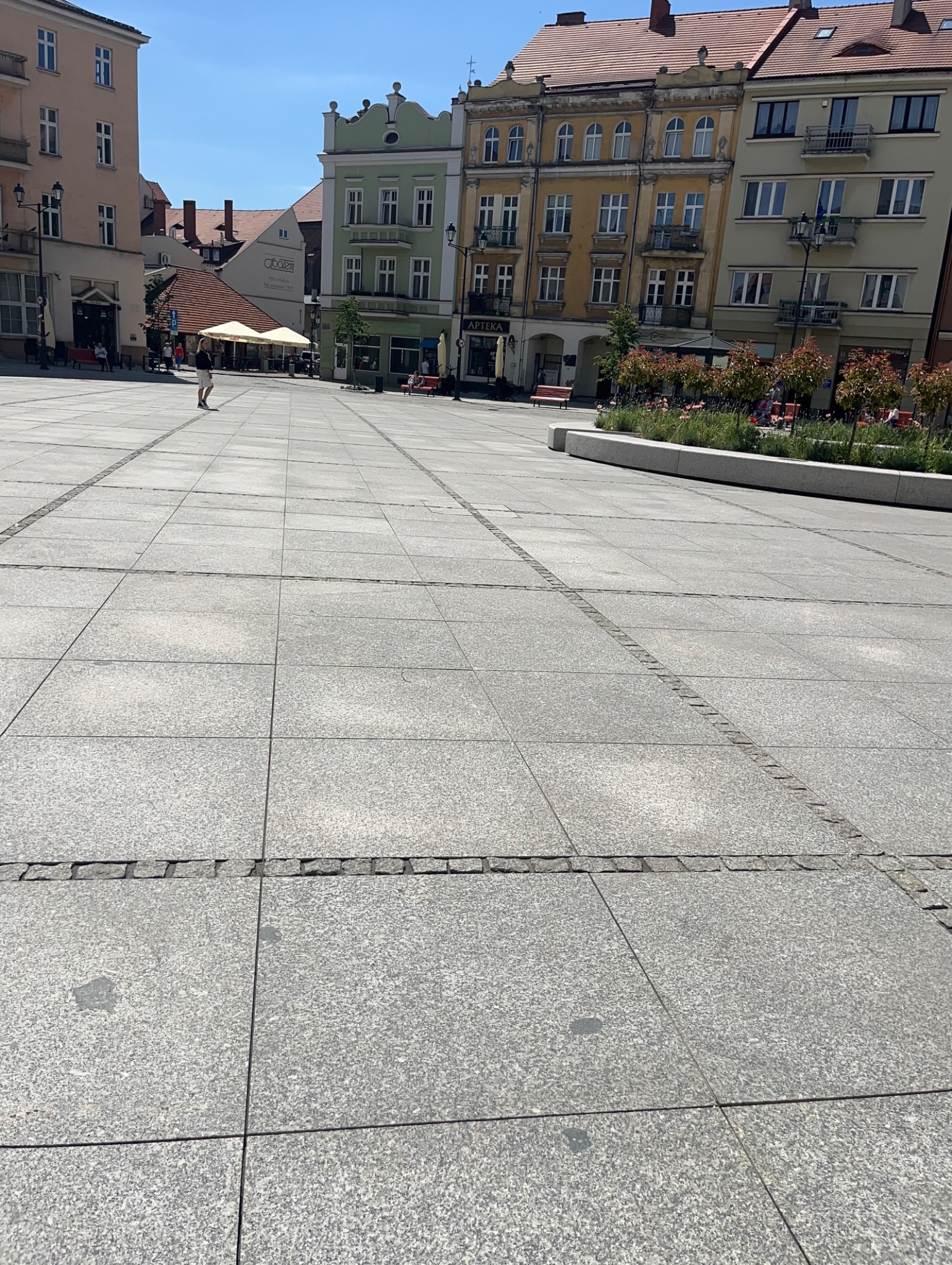 Rozległy plac miejski wyłożony szarą kostką brukową, z widocznymi liniami z drobnej kostki oddzielającymi poszczególne sekcje, w tle zabytkowe budynki i elementy zieleni miejskiej.