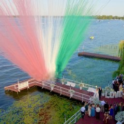 Piotr Wołowik Fotografia - Ślub nad jeziorem: para młoda na pomoście podczas wystrzału kolorowych dymów w barwach włoskiej flagi, otoczona gośćmi i dekoracją LOVE z kwiatów.