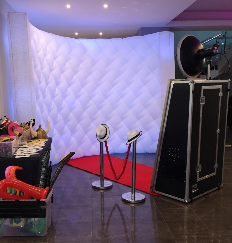 Fotolustro z rekwizytami i czerwonym dywanem w Lubartowie, ustawione przed białą, pikowaną ścianką. Dwa kapelusze na stojakach wyznaczają wejście.