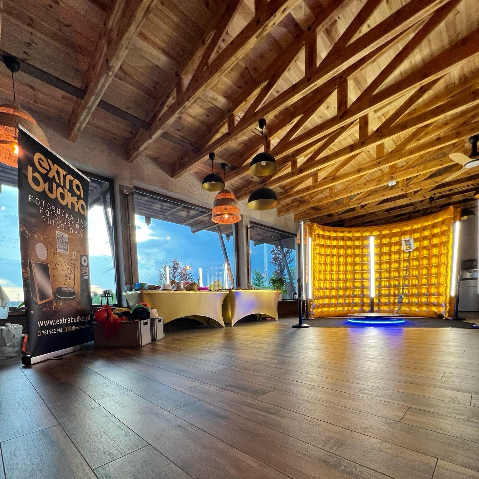 Sala z drewnianym stropem, przygotowana do imprezy z żółtą dmuchaną ścianką i obrotową platformą z niebieskim oświetleniem, widoczny baner reklamowy.