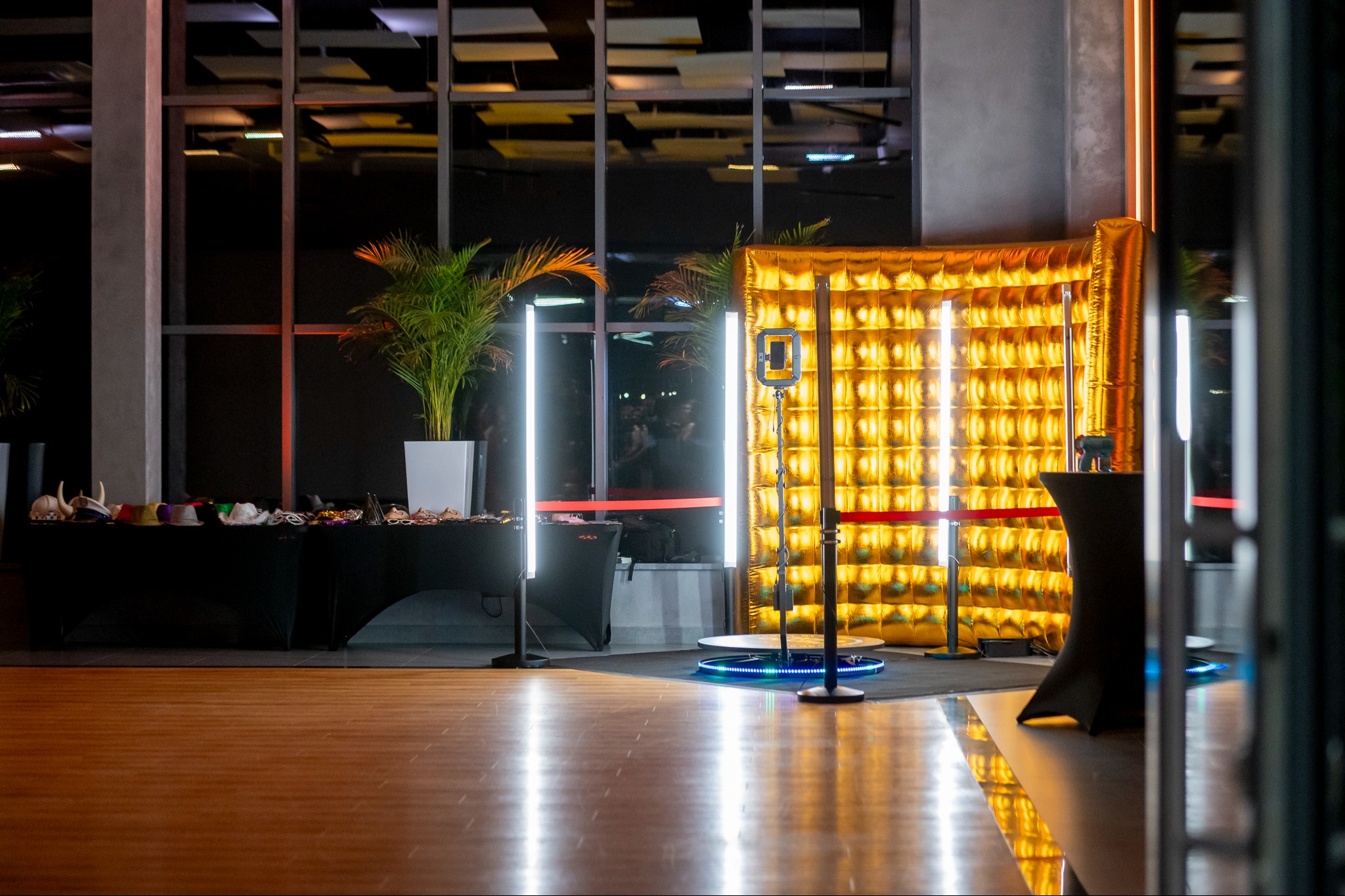 Złota, dmuchana ścianka z kratownicą, oświetlona lampami LED, z platformą obrotową i stolikiem z czarnym obrusem, w tle stół z rekwizytami do zdjęć i rośliny doniczkowe.