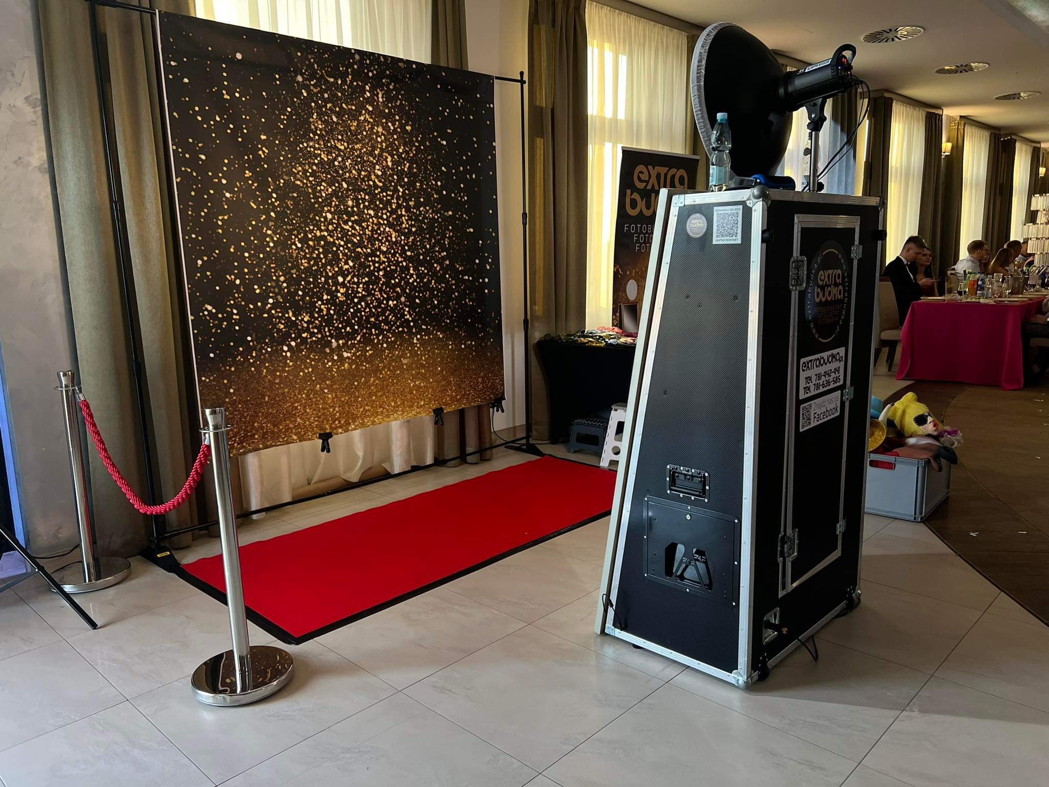 Fotolustro z czarną obudową, oświetleniem i czerwonym dywanem na tle błyszczącej ścianki, ustawione w sali bankietowej.