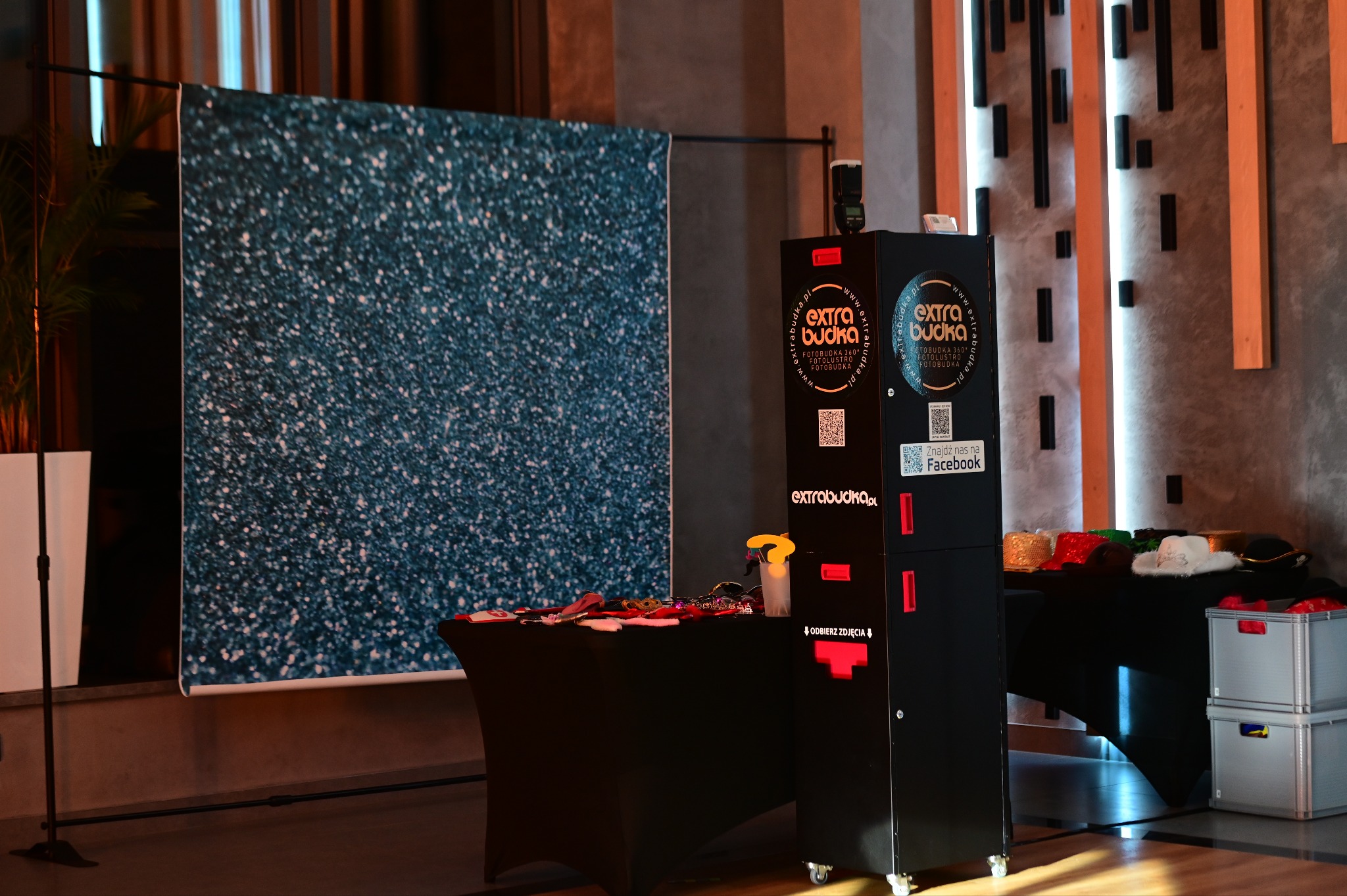 Kompletna konfiguracja mobilnej atrakcji na imprezy: czarna obudowa urządzenia z logo i ekranem, tło z efektem brokatu, stolik z rekwizytami do zdjęć, całość ustawiona w pomieszczeniu z elementami...