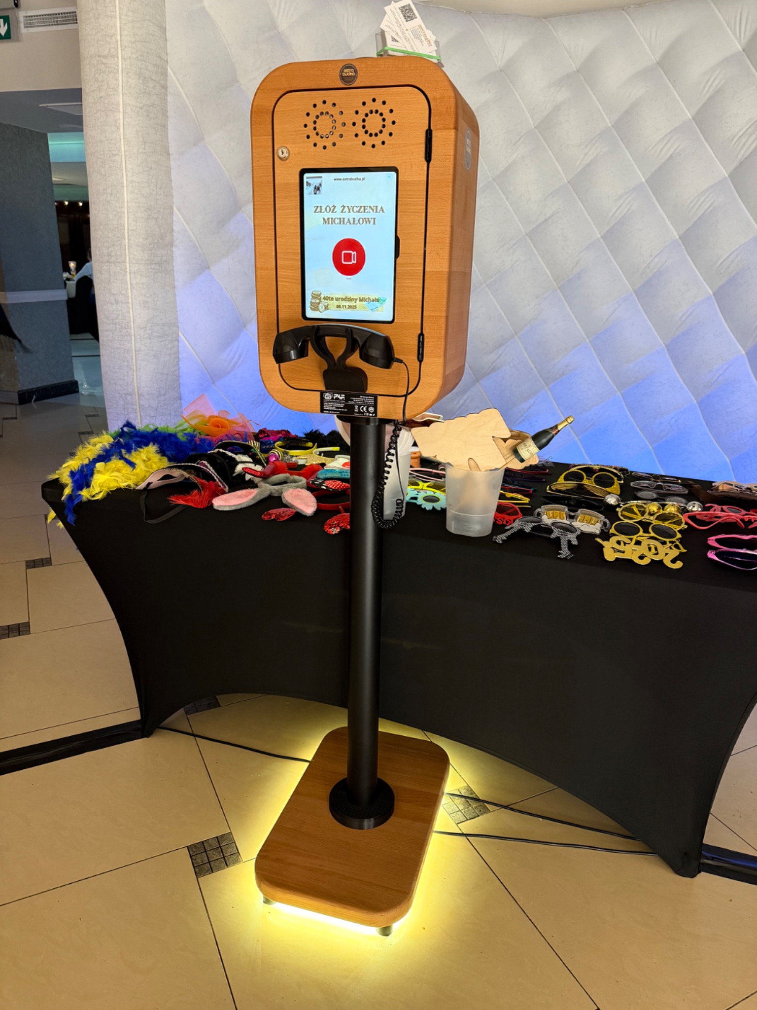 Drewniana fotobudka z ekranem dotykowym i rekwizytami na stole, gotowa do użycia na imprezie. Podświetlana podstawa dodaje elegancji.