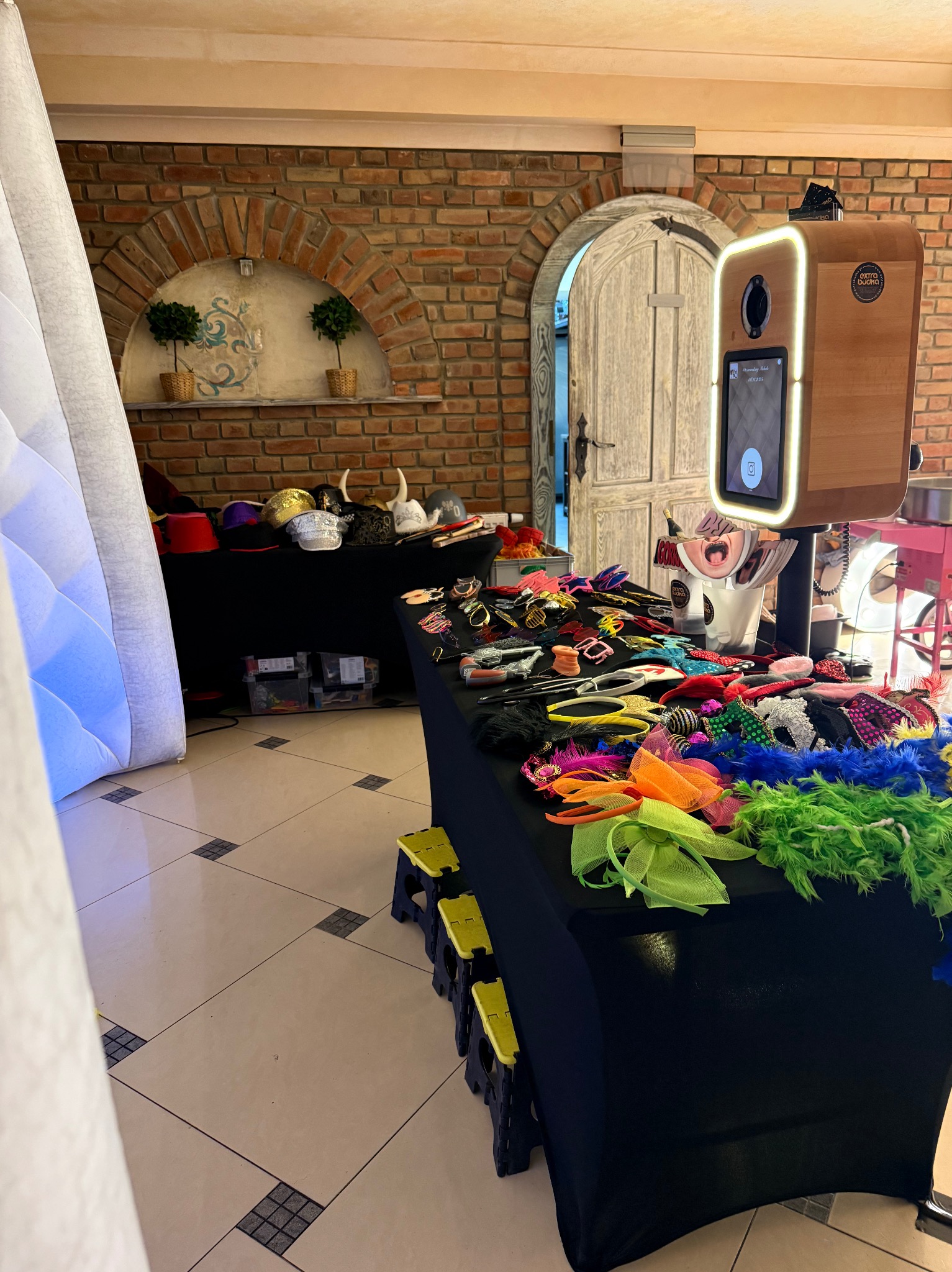 Fotobudka z rekwizytami na stole: kolorowe boa, maski, kapelusze. W tle ceglana ściana i dekoracyjne elementy. Gotowe do zabawy na imprezie.