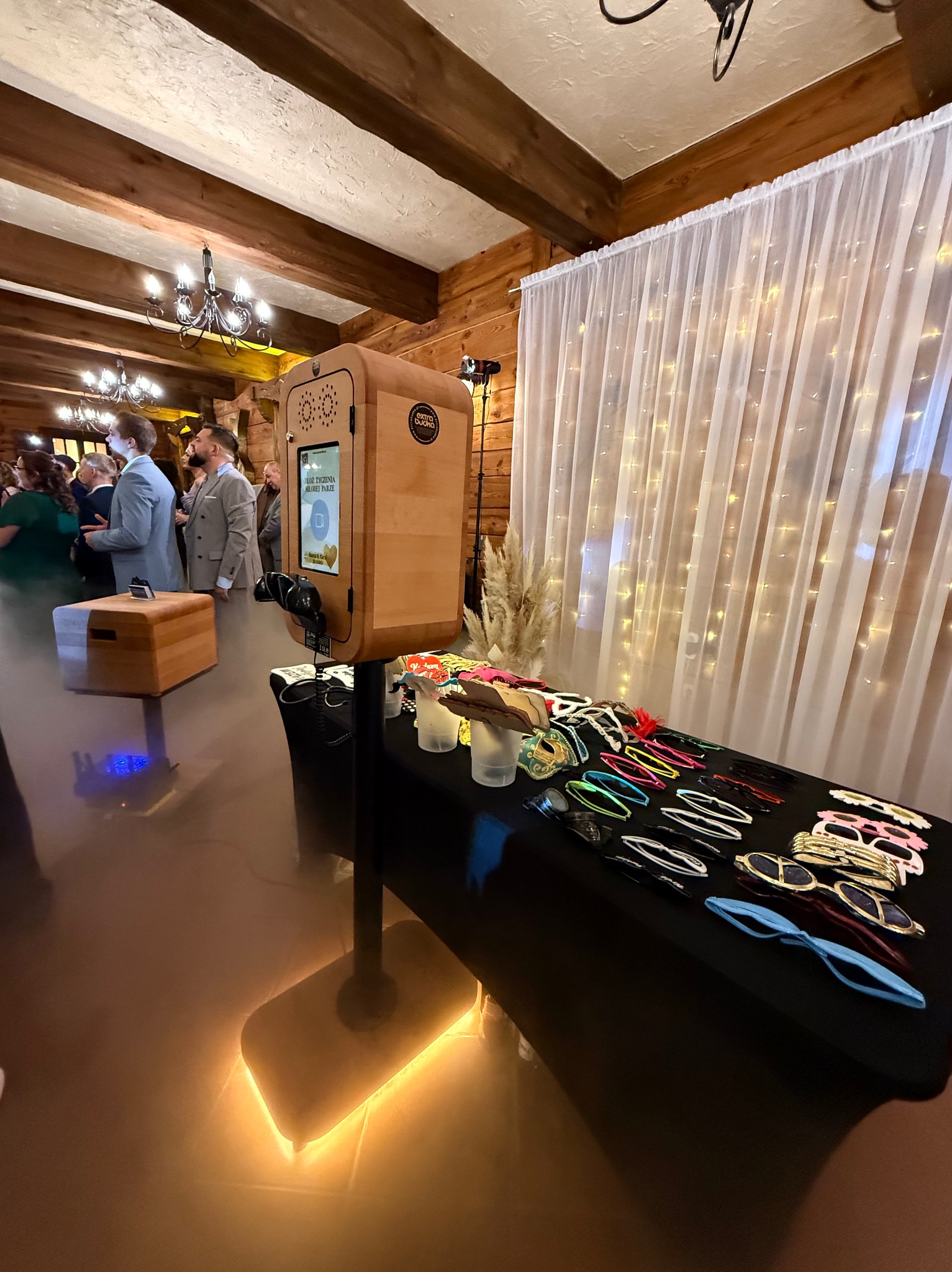 Drewniana fotobudka z ekranem dotykowym na tle gości weselnych i białej, świetlnej kurtyny. Na stole leżą rekwizyty: okulary i maski.