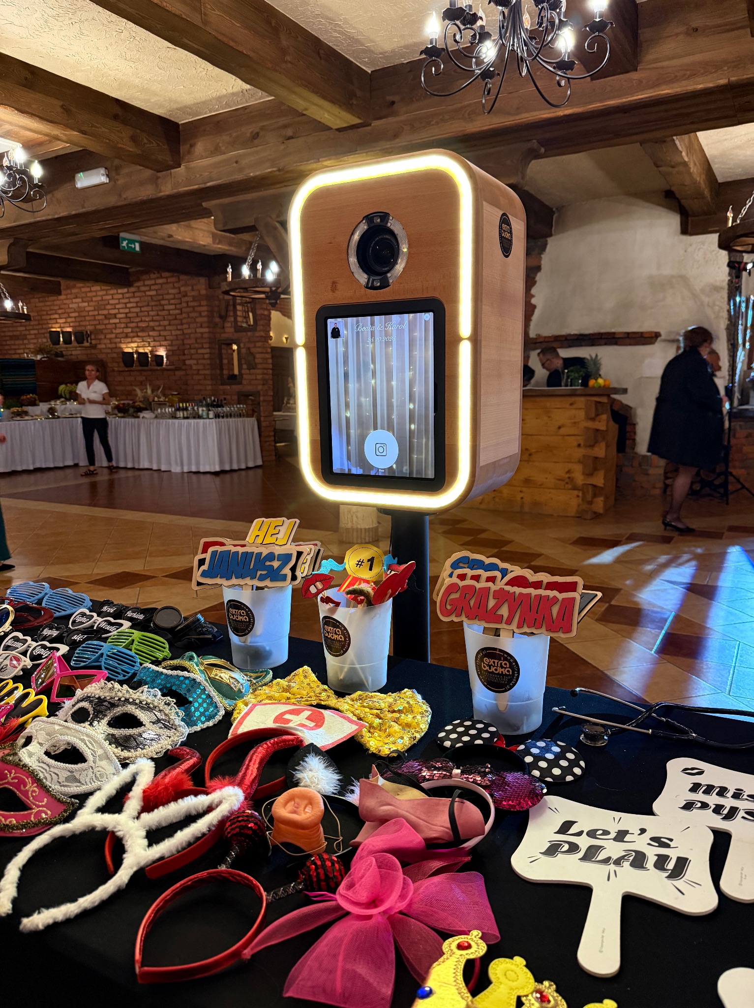 Fotobudka z rekwizytami: maski, okulary, tabliczki z imionami 'Janusz' i 'Grażynka'. Dekoracje na stole, w tle sala bankietowa z żyrandolami.