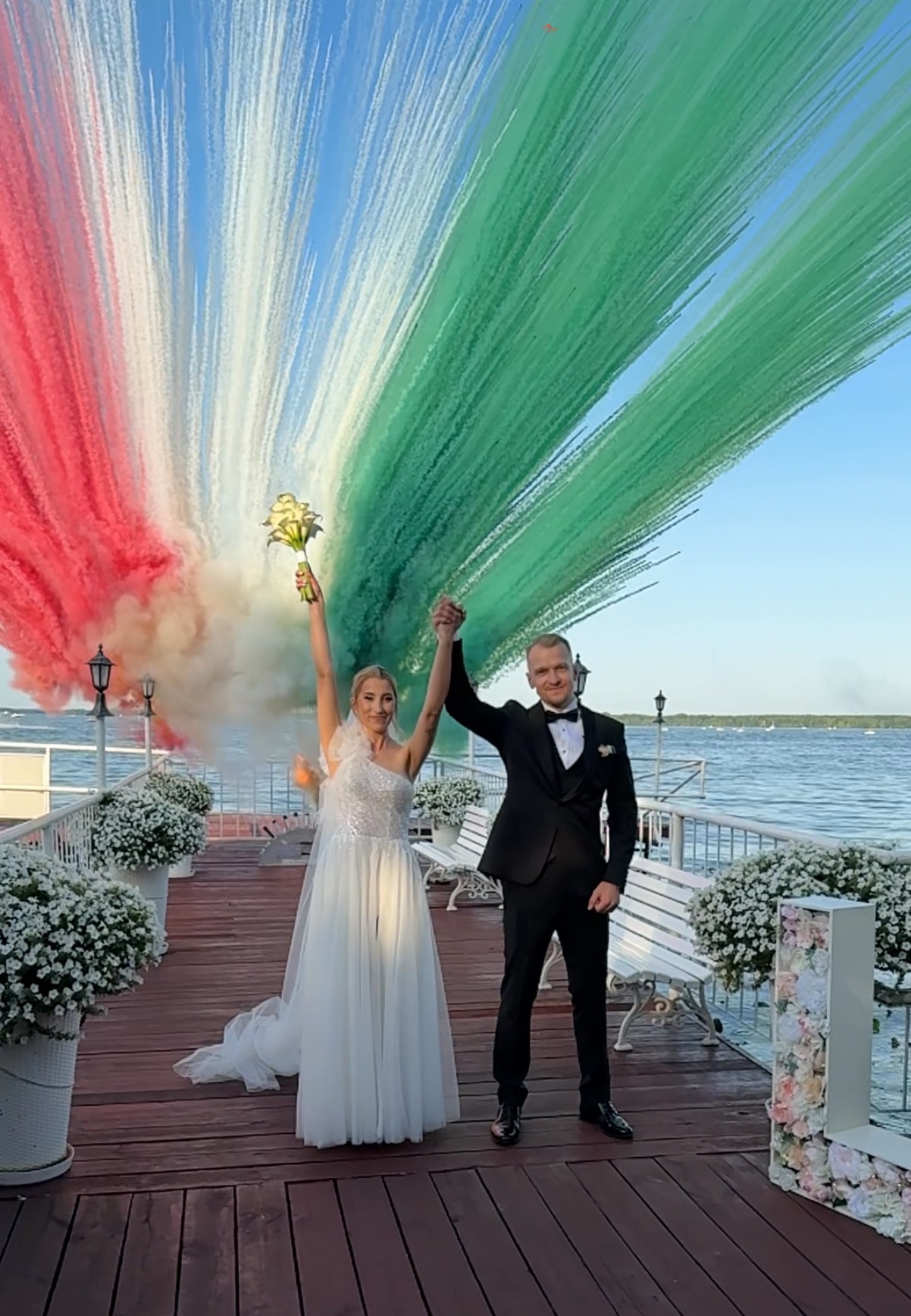 Para młoda na molo, świętująca z kolorowymi pióropuszami dymu w barwach flagi Włoch. Ona w białej sukni, on w smokingu. W tle woda i dekoracje kwiatowe.