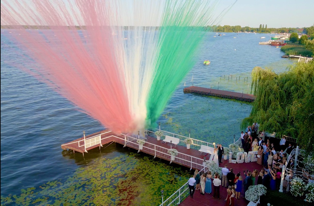 Ślub nad jeziorem: para młoda na pomoście podczas wystrzału kolorowych dymów w barwach włoskiej flagi, otoczona gośćmi i dekoracją LOVE z kwiatów.