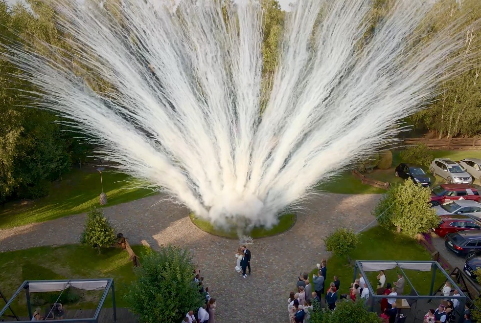 Para młoda w otoczeniu gości i efektownej fontanny iskier podczas ceremonii ślubnej, uchwycone z lotu ptaka. Widoczne drzewa, zaparkowane samochody i dekoracje.