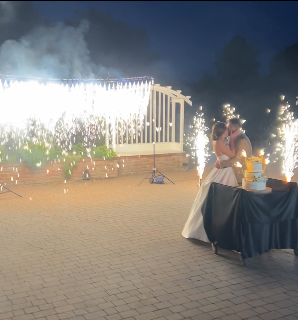 Pocałunek pary młodej przy torcie weselnym, w tle fontanny iskier i biała altana. Ujęcie z niskiego kąta, wieczorne oświetlenie.