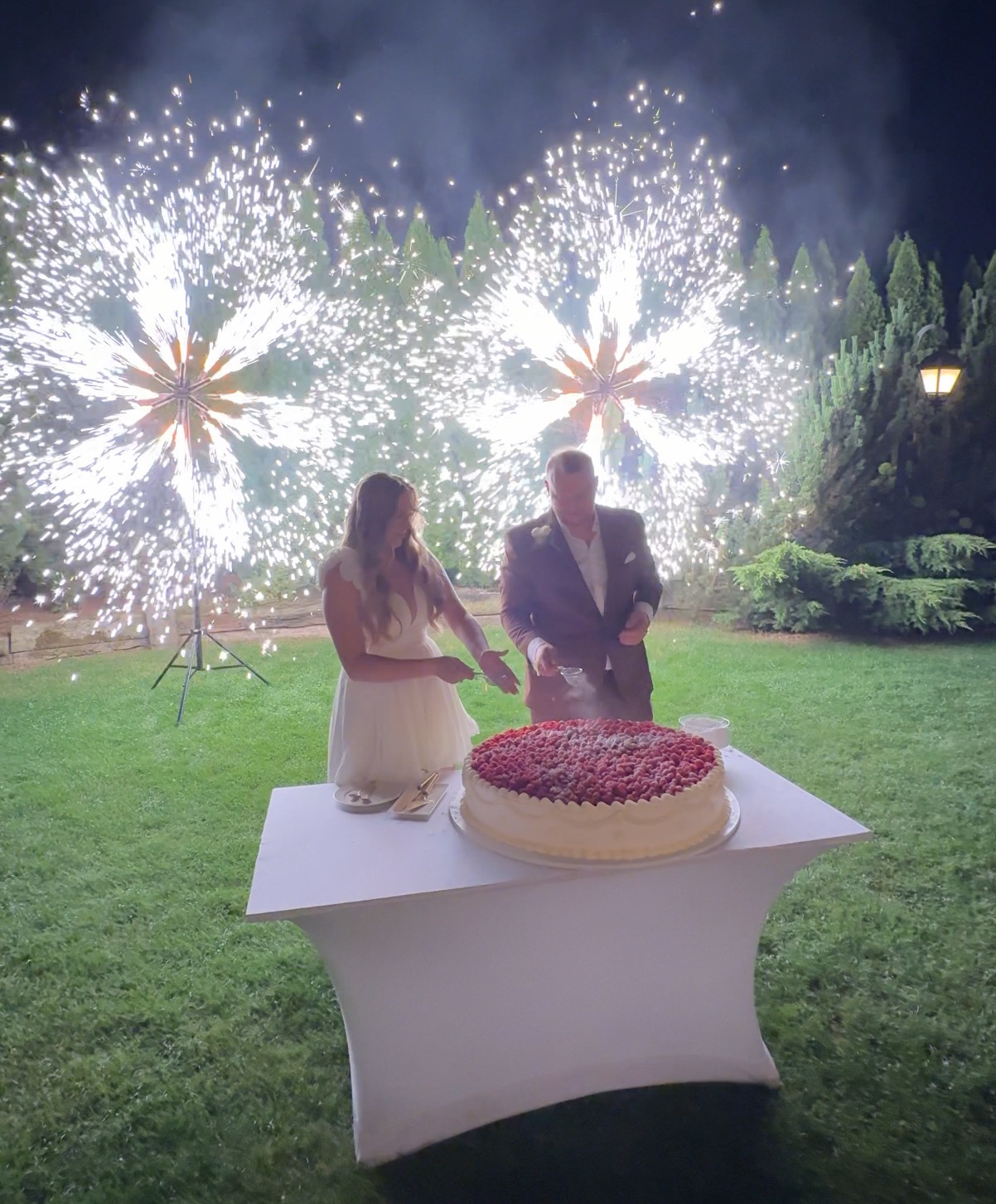 Para młoda kroi tort weselny na tle efektownych fontann iskier podczas nocnej uroczystości plenerowej. Romantyczna atmosfera.