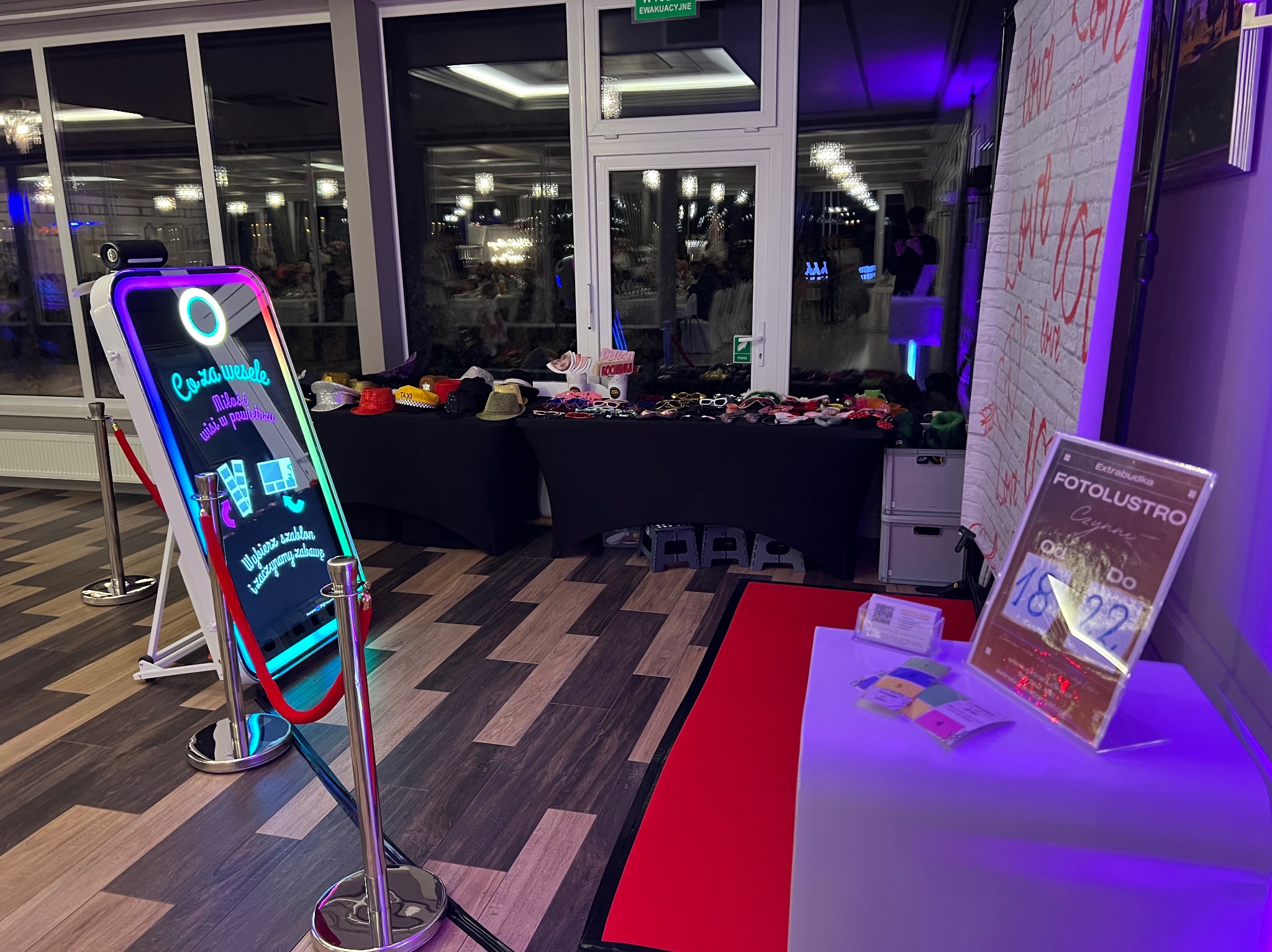 Fotobudka z ekranem dotykowym i rekwizytami na stole, gotowa do użycia na imprezie. W tle widoczni goście i dekoracje sali weselnej.