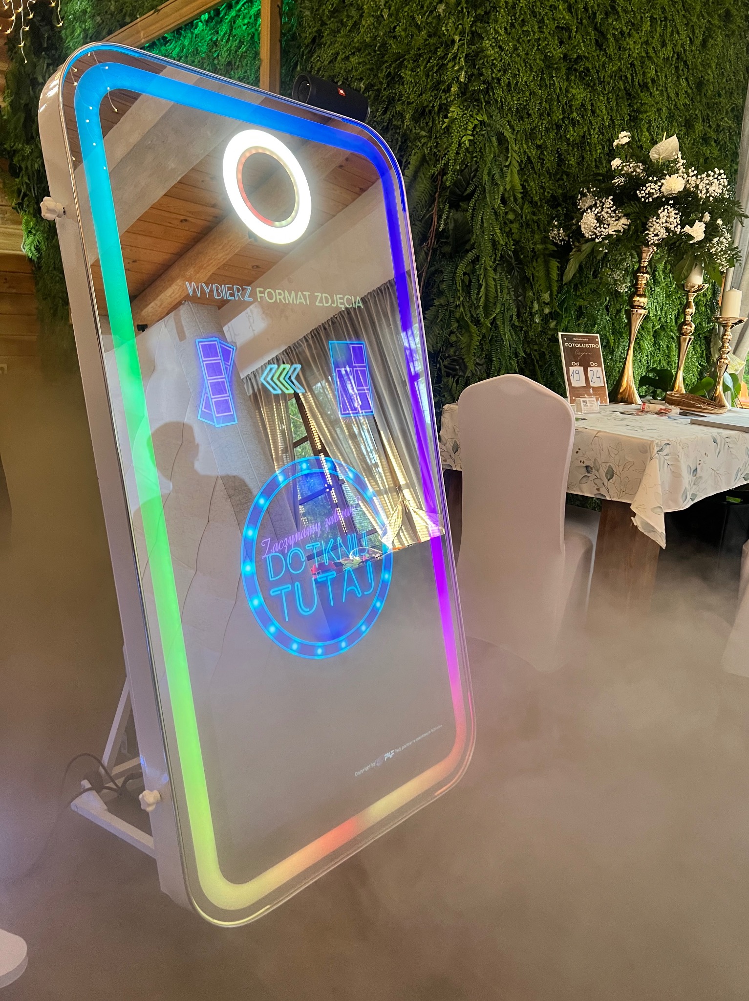 Interaktywna fotobudka lustro z neonowym obramowaniem na tle zielonej ściany i dekoracji kwiatowych, otoczona subtelną mgłą, gotowa do uwieczniania radosnych chwil.