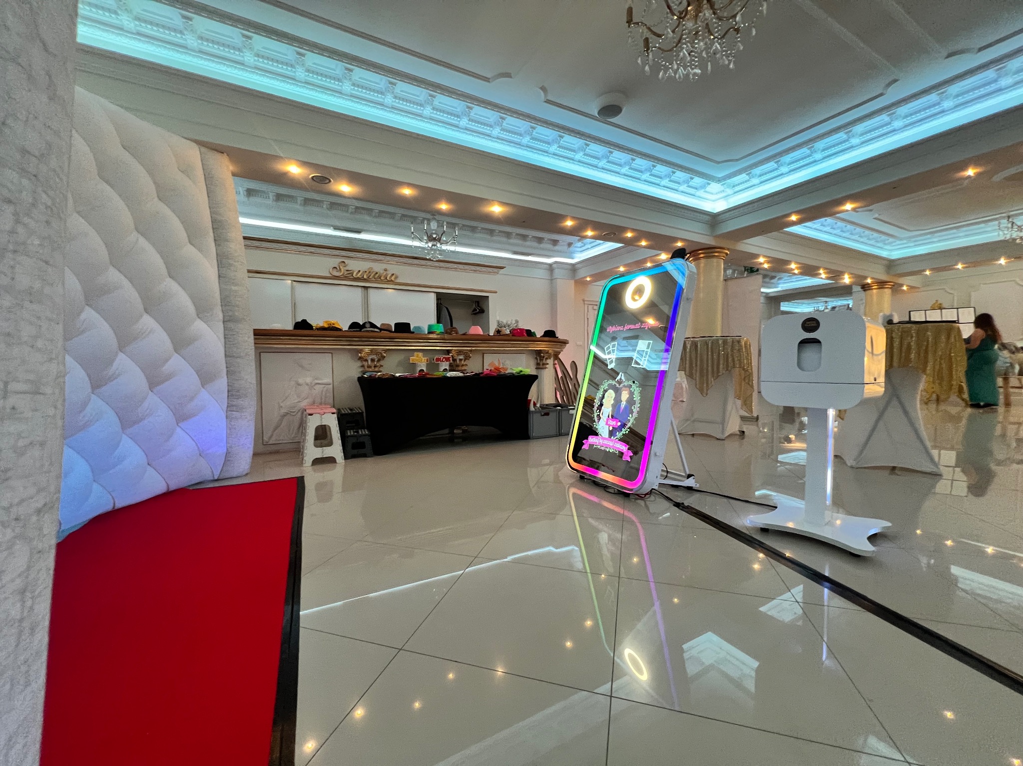Elegancka fotobudka z neonowym obramowaniem na tle sali bankietowej z dekoracjami i rekwizytami. Widoczny fragment dmuchanej ścianki i błyszcząca podłoga.