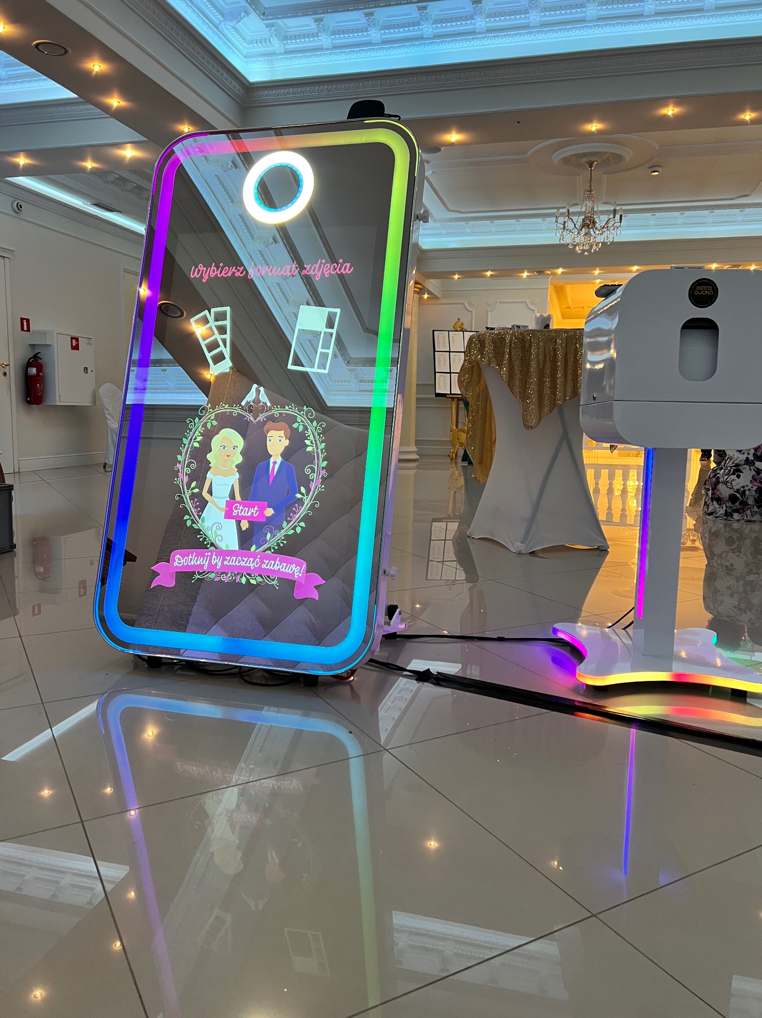 Interaktywna fotobudka lustrzana z animacją pary młodej i napisem 'Dotknij by zacząć zabawę!' na błyszczącej podłodze sali balowej.