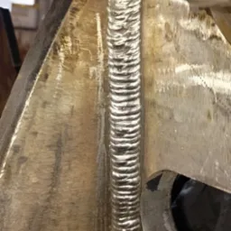 Szczegółowe zbliżenie na spawany element konstrukcyjny z metalu, ukazujące precyzyjne i równe spoiny wzdłuż krawędzi, z widocznymi śladami obróbki metalu i niedoskonałościami powierzchni.