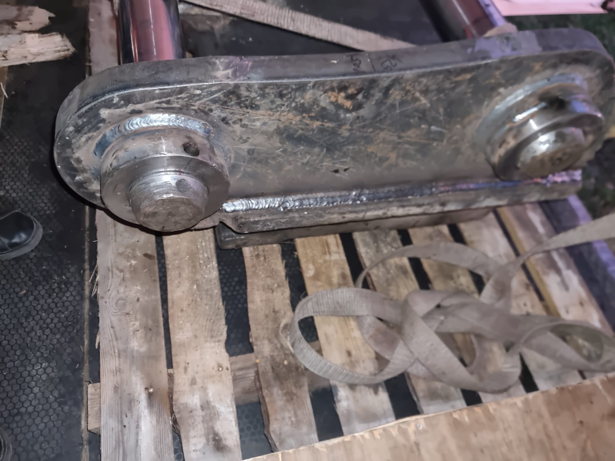Ciężki, metalowy element konstrukcyjny z widocznymi śladami spawania, leżący na drewnianej palecie, z fragmentem czarnego buta widocznym w lewym dolnym rogu.