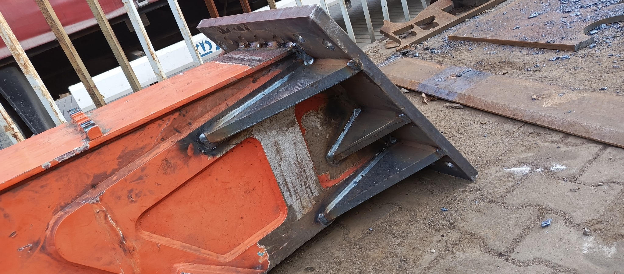 Pomarańczowa konstrukcja stalowa z widocznymi spawami, leżąca na ziemi, fragment ogrodzenia w tle.