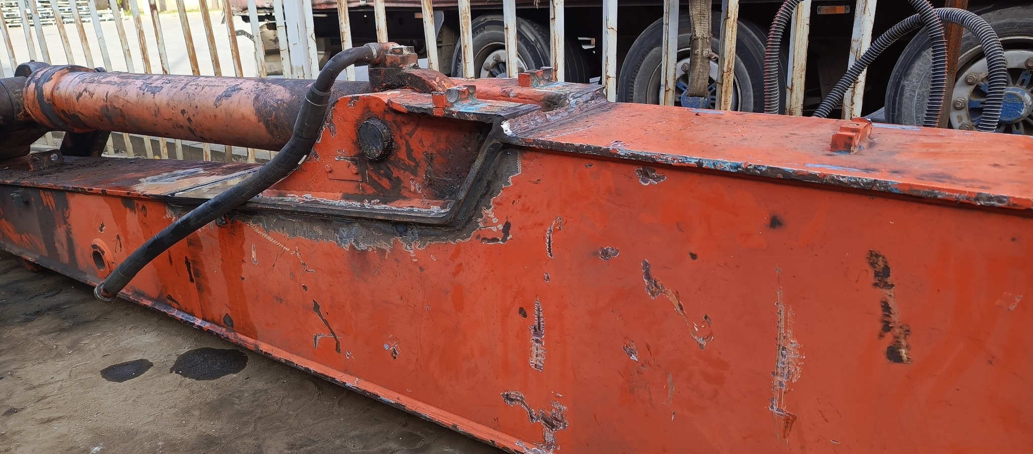 Pomarańczowy element konstrukcyjny z widocznymi śladami użytkowania, rdzą i odpryskami farby, prawdopodobnie część maszyny budowlanej lub przemysłowej, z przewodami hydraulicznymi i elementami...